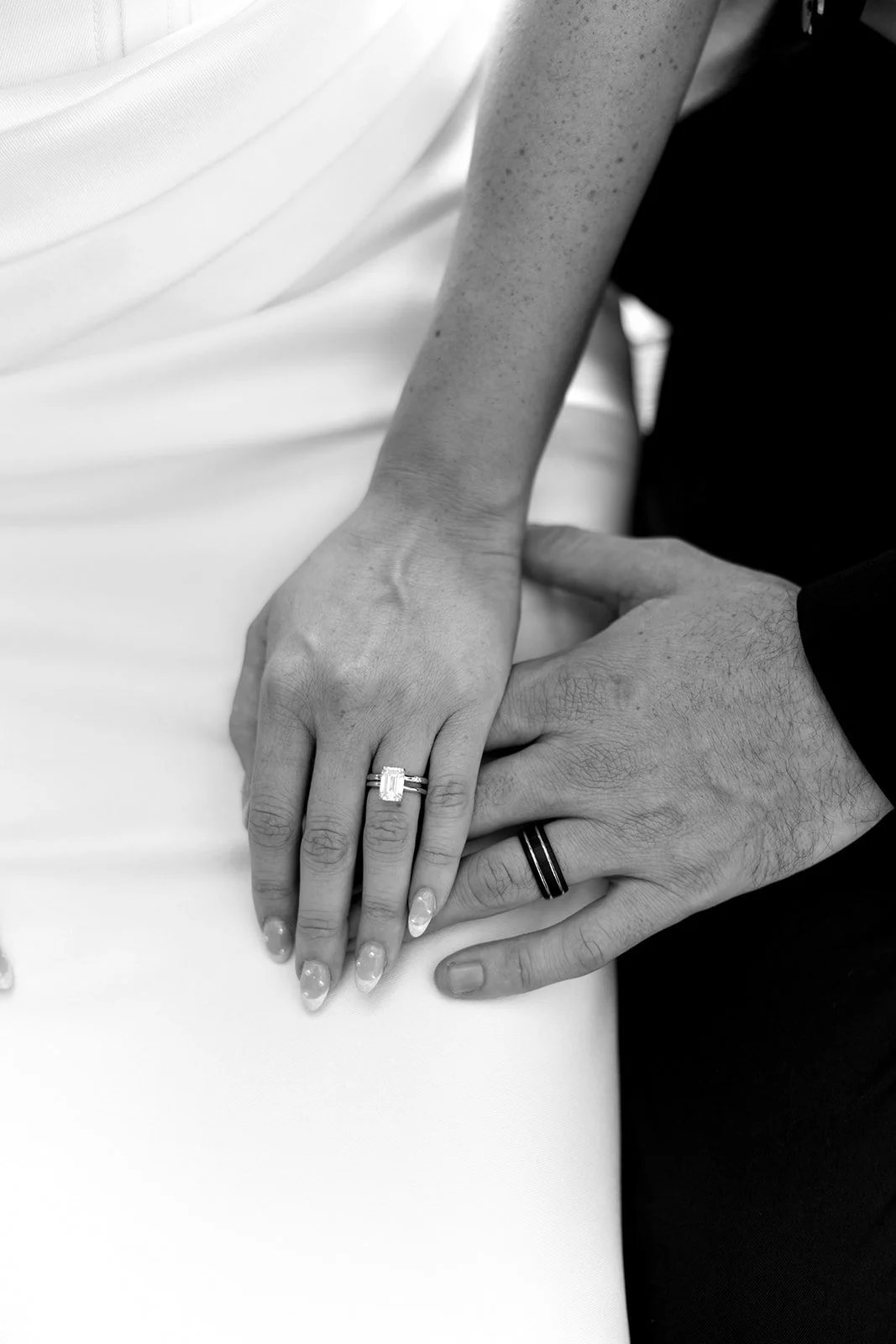 Close-up of a woman's hand with a wedding ring, resting on a man's hand with a wedding band, both hands on a white surface.