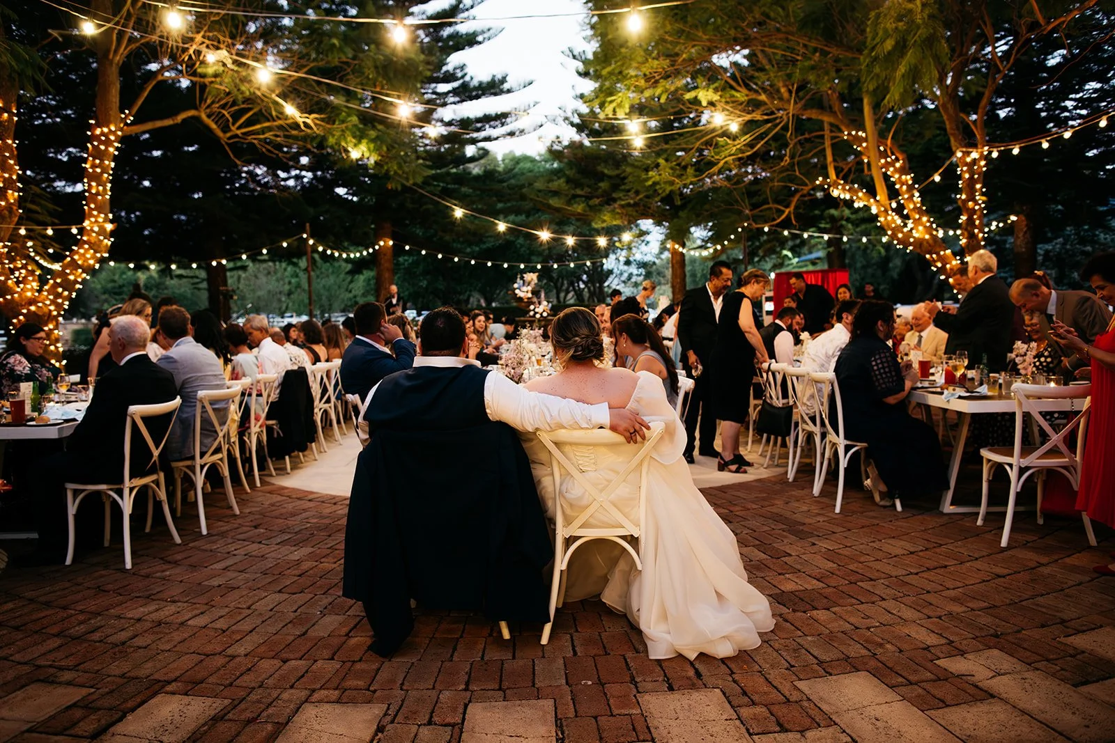 Outdoor wedding reception with string lights, bridal couple sitting at center, surrounded by guests at tables, trees decorated with fairy lights, romantic evening ambiance.