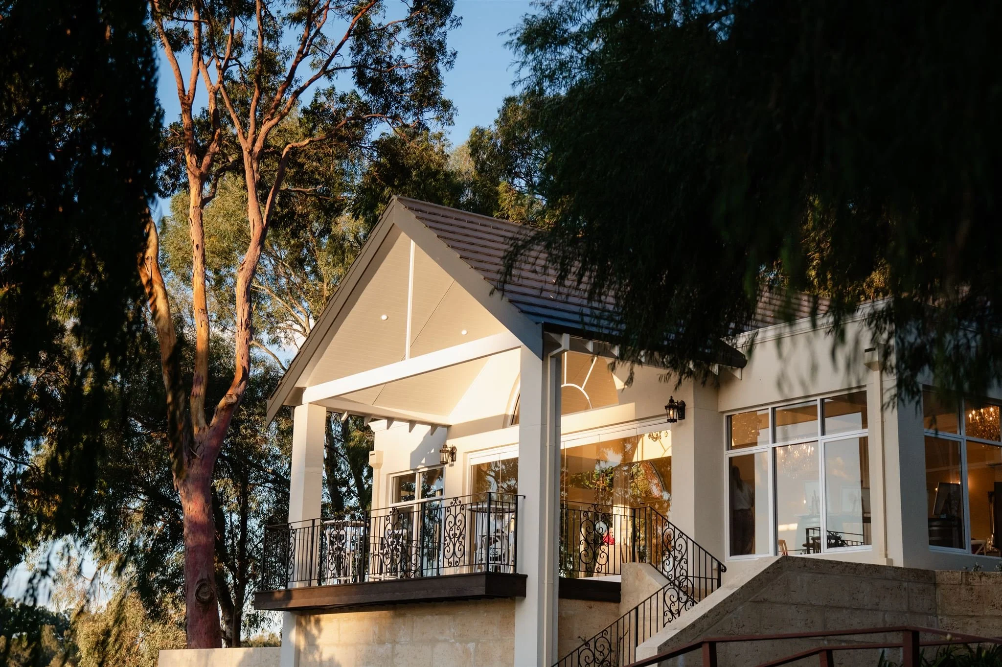 A modern house with large windows and a balcony, surrounded by tall trees, under a clear blue sky.