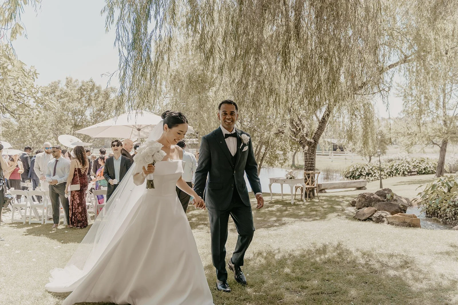 Bride and groom walking hand in hand outdoors at a wedding reception, with guests in the background under umbrellas.