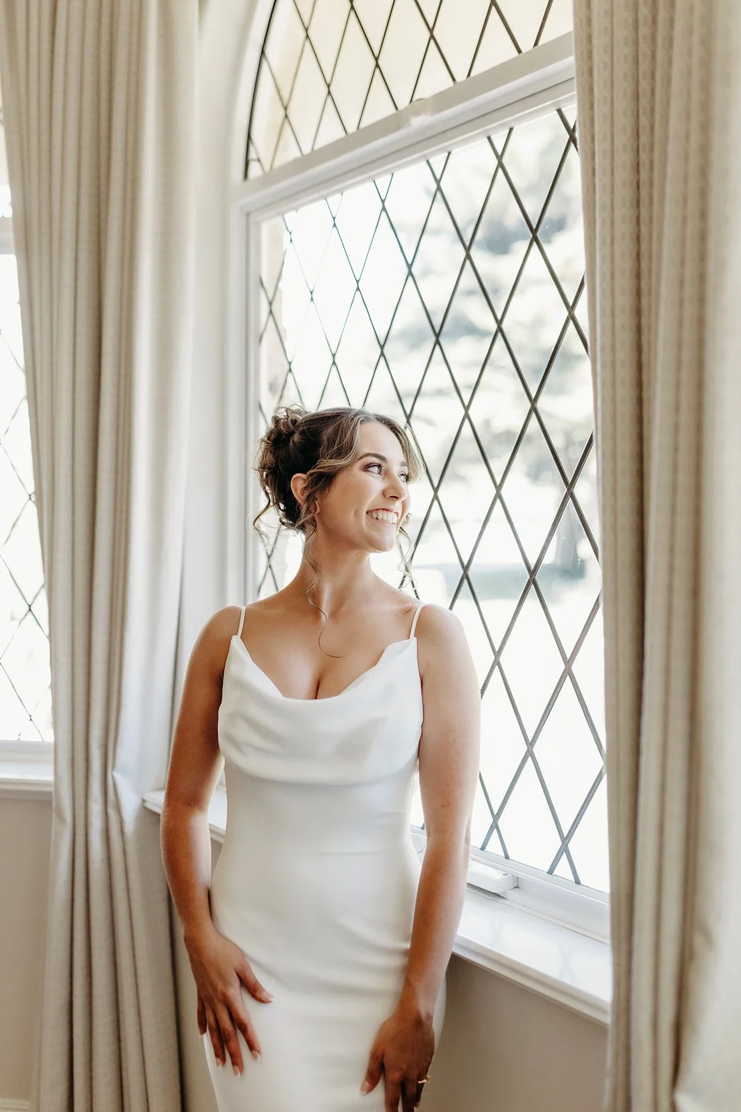A bride in a white wedding dress standing by a window with diamond-shaped leaded glass, smiling and looking outside.