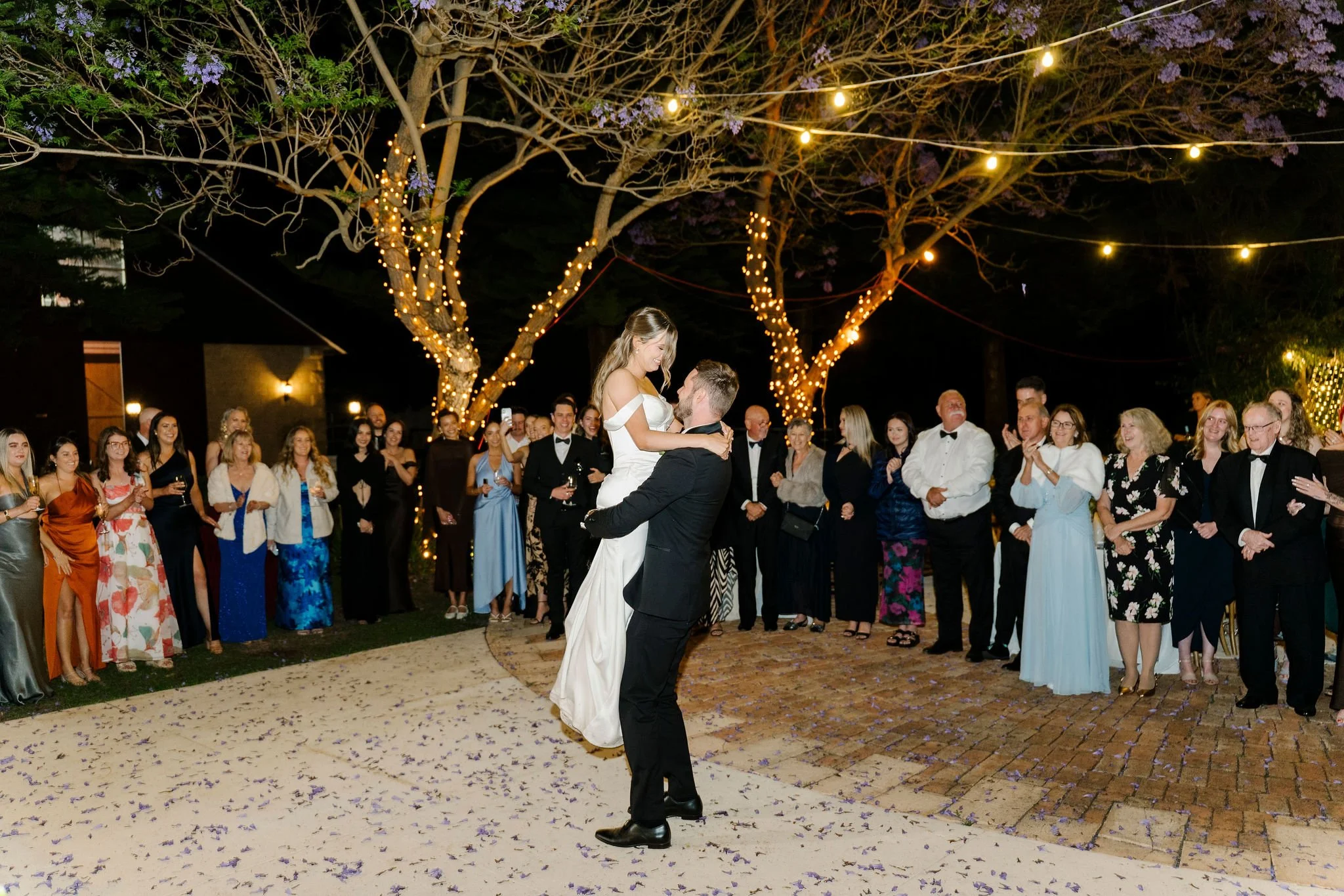A bride and groom dancing at their wedding reception outdoors at night, surrounded by guests. The bride wears a white gown, and the groom is in a black suit. String lights and fairy lights are hung around trees, creating a festive atmosphere.