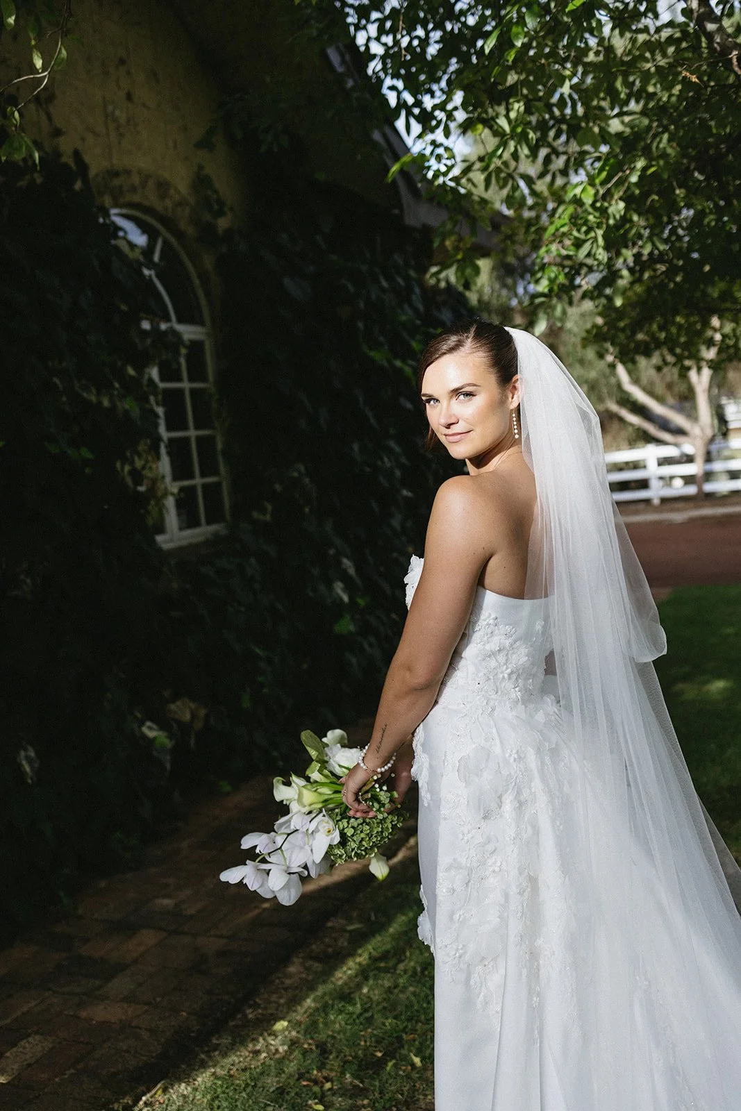 Bride in a white wedding dress and veil holding a bouquet of white flowers outdoors.