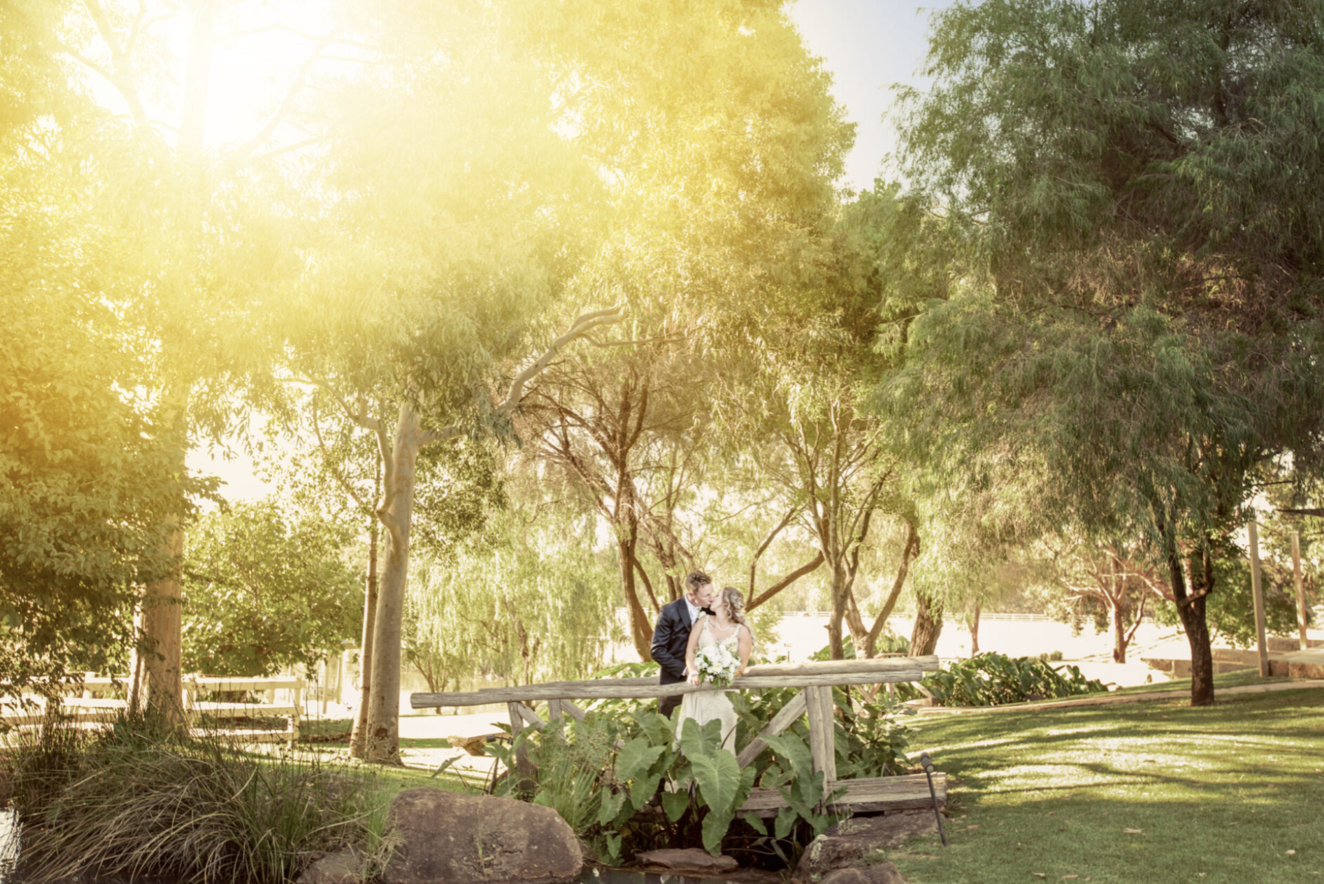 A bride and groom sharing a kiss on a park bridge with trees and greenery around them, during a sunny day.