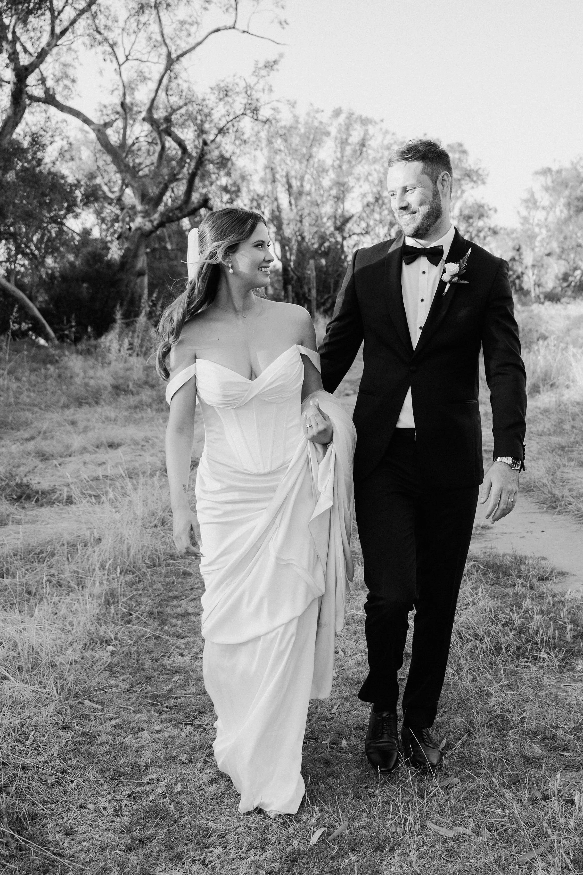 A bride and groom walking outdoors, smiling at each other on their wedding day in a natural setting.
