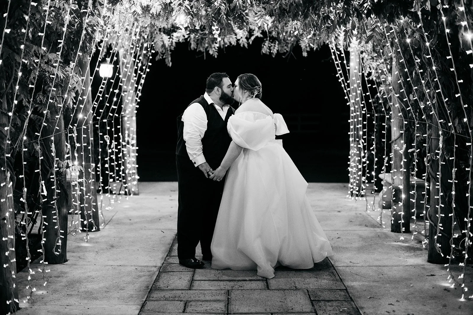 A couple sharing a kiss during their wedding under a decorated archway with string lights at night.