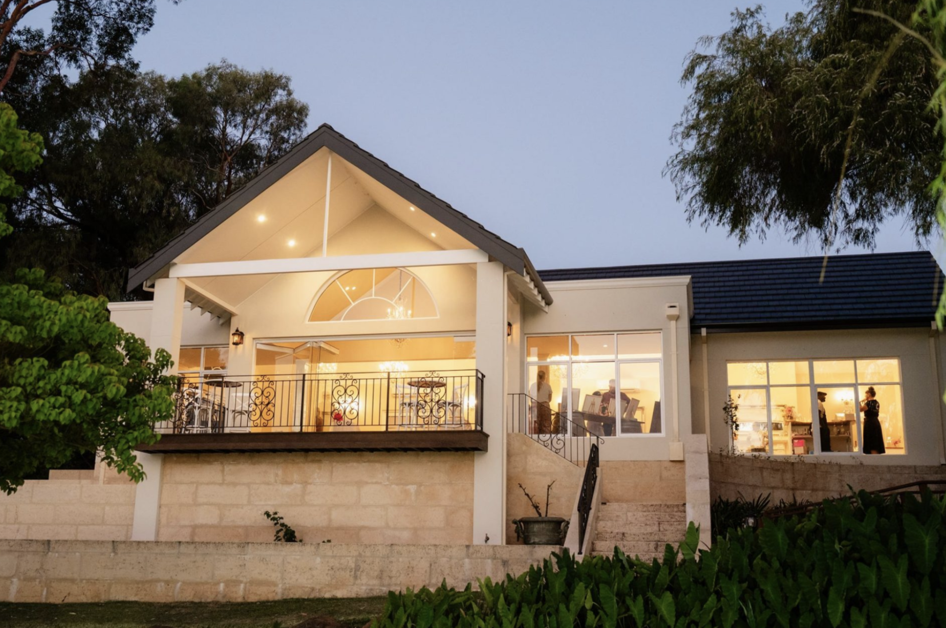 A two-story house at dusk with an illuminated interior, a balcony, large windows, surrounded by trees and greenery.