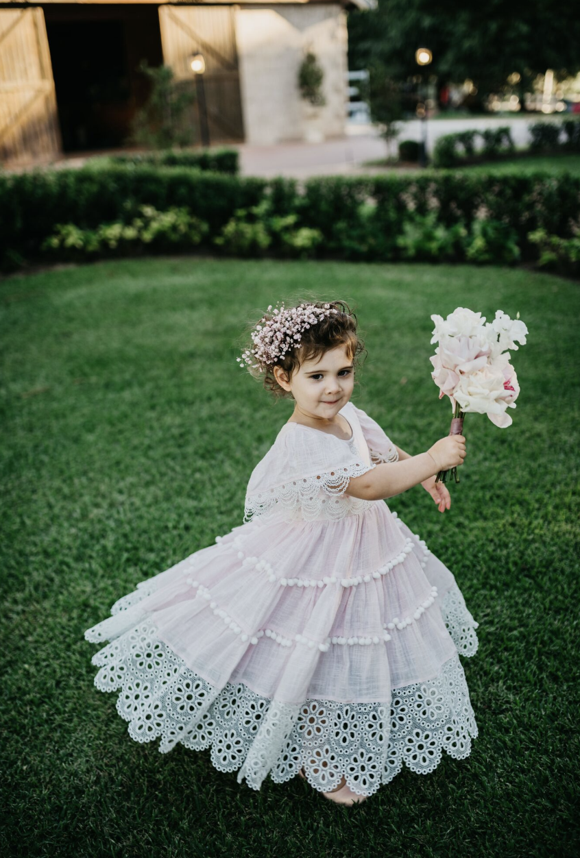 A young girl in a pink dress with lace and embroidered details, holding a bouquet of flowers, standing on a well-manicured lawn with bushes and a building in the background.