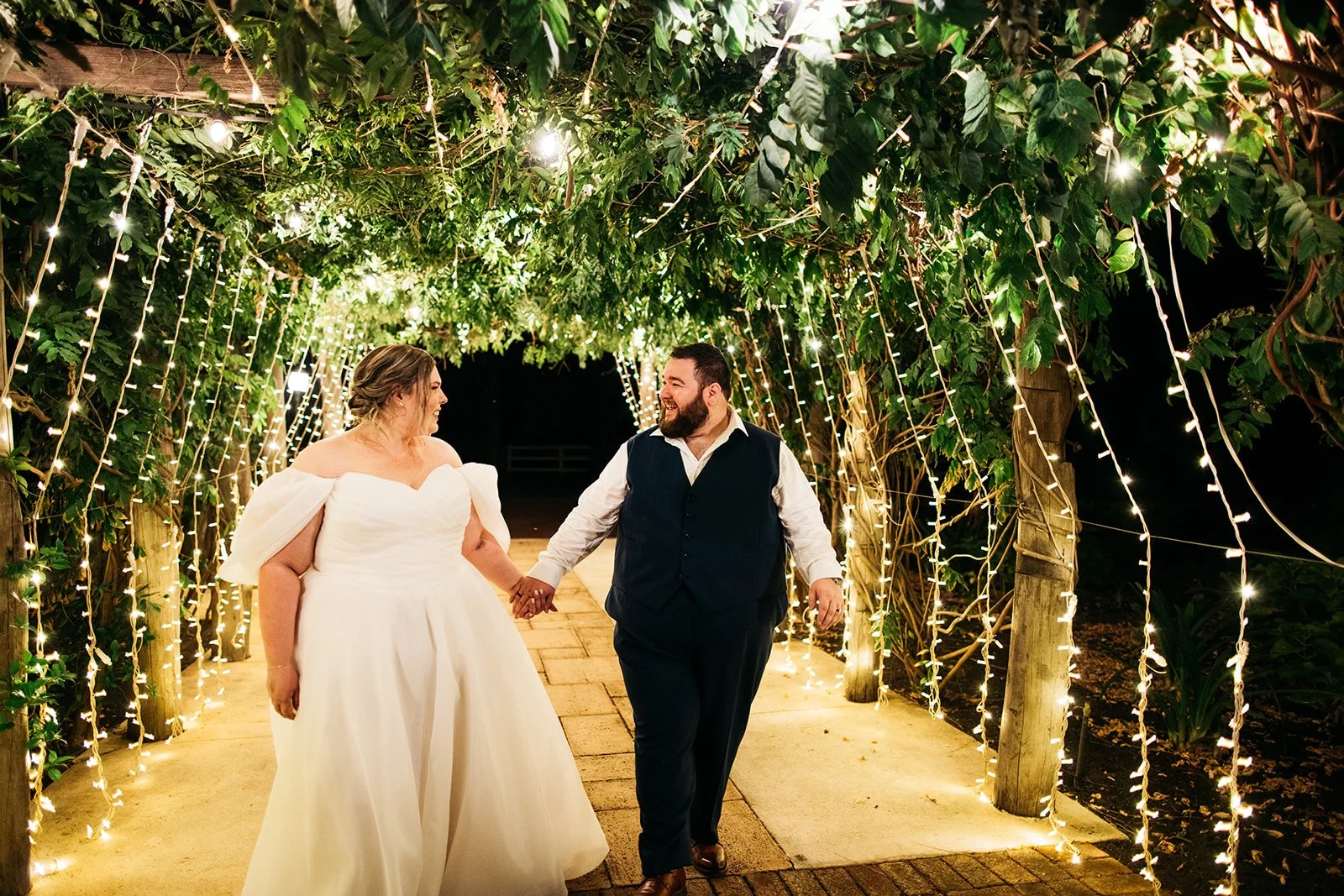 A newlywed couple walking hand in hand through a tunnel of green foliage decorated with string lights during nighttime.