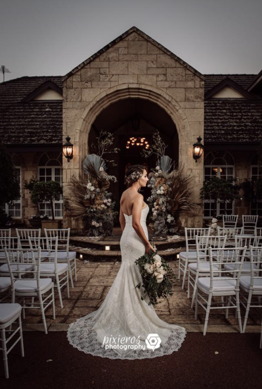 A bride in a strapless white wedding dress holding a bouquet of flowers, standing outdoors in front of a church with stone walls and arched entrance, decorated with floral arrangements and outdoor lighting, under a cloudy sky.