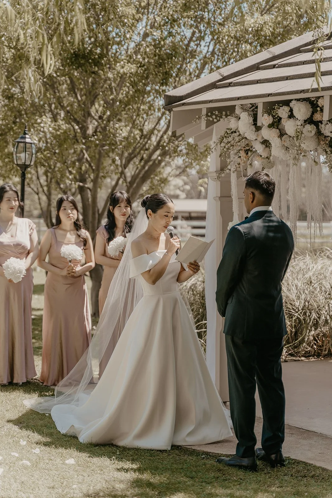 Bride reading vows at outdoor wedding ceremony with groom and bridesmaids in background.
