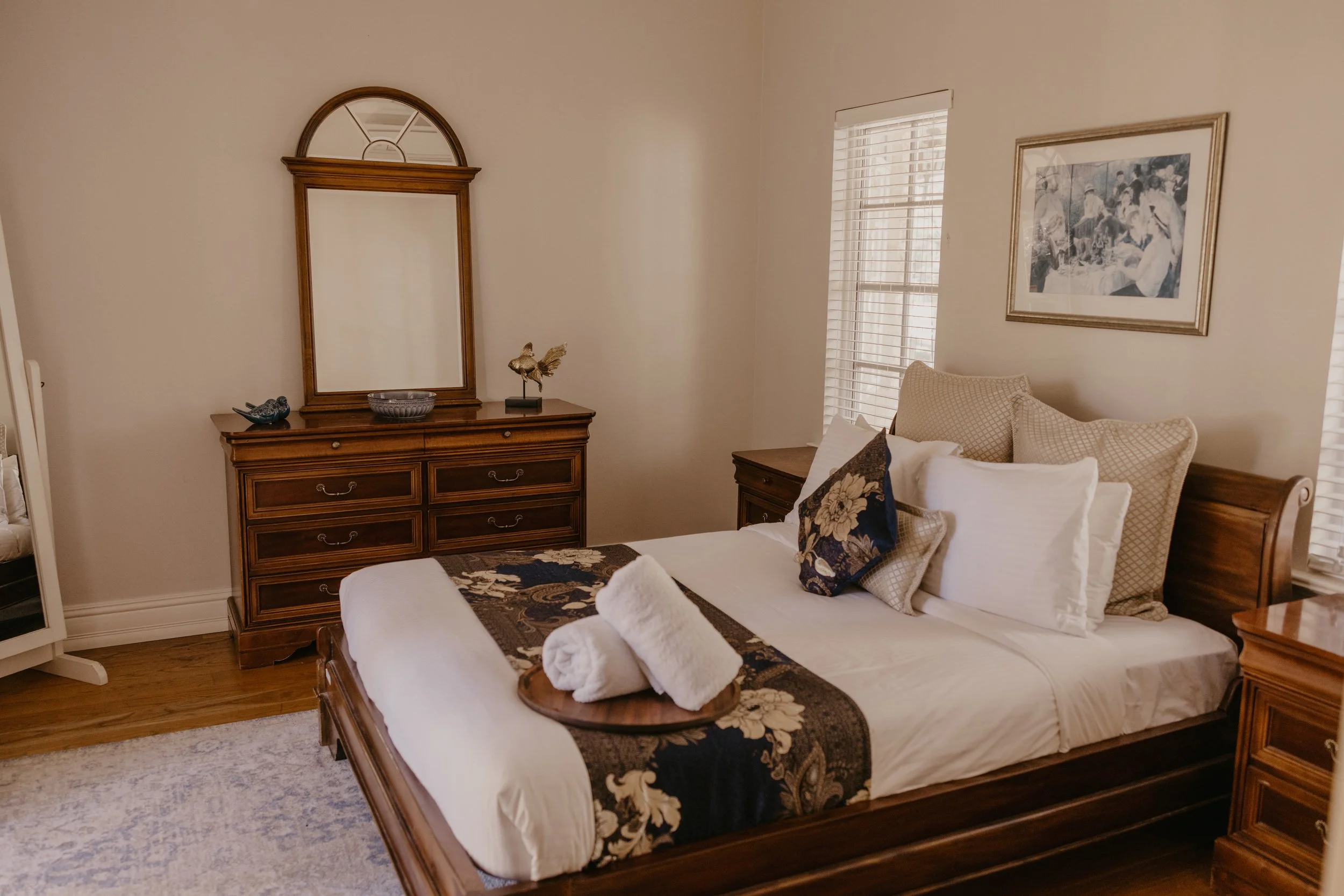 A bedroom with a wooden bed frame, white bedding, and decorative pillows, next to a wooden dresser with a mirror, a shell-shaped dish, a bird figurine, and a decorative bowl, with a window with blinds and a framed picture on the wall.