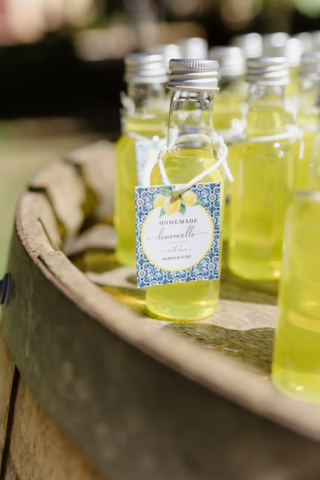 Small glass bottles filled with homemade limoncello, labeled with a decorative tag featuring lemons, placed on a wooden surface outdoors.