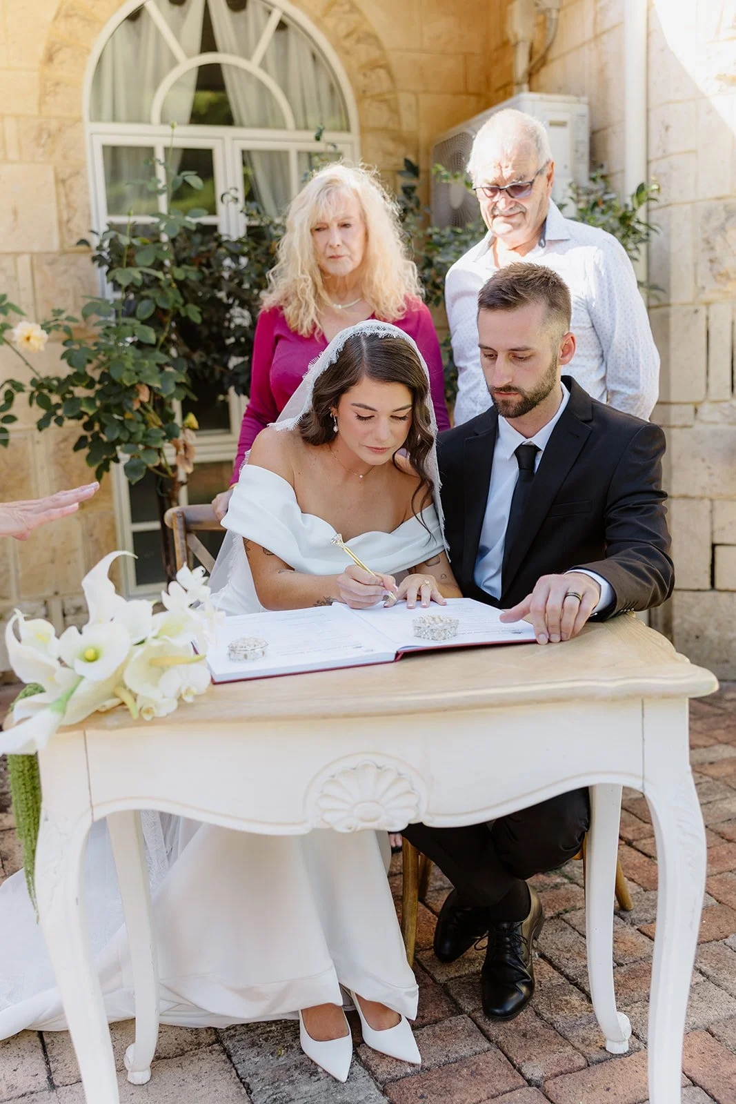 Bride and groom signing marriage documents outdoors with two witnesses and a floral arrangement on a white table.