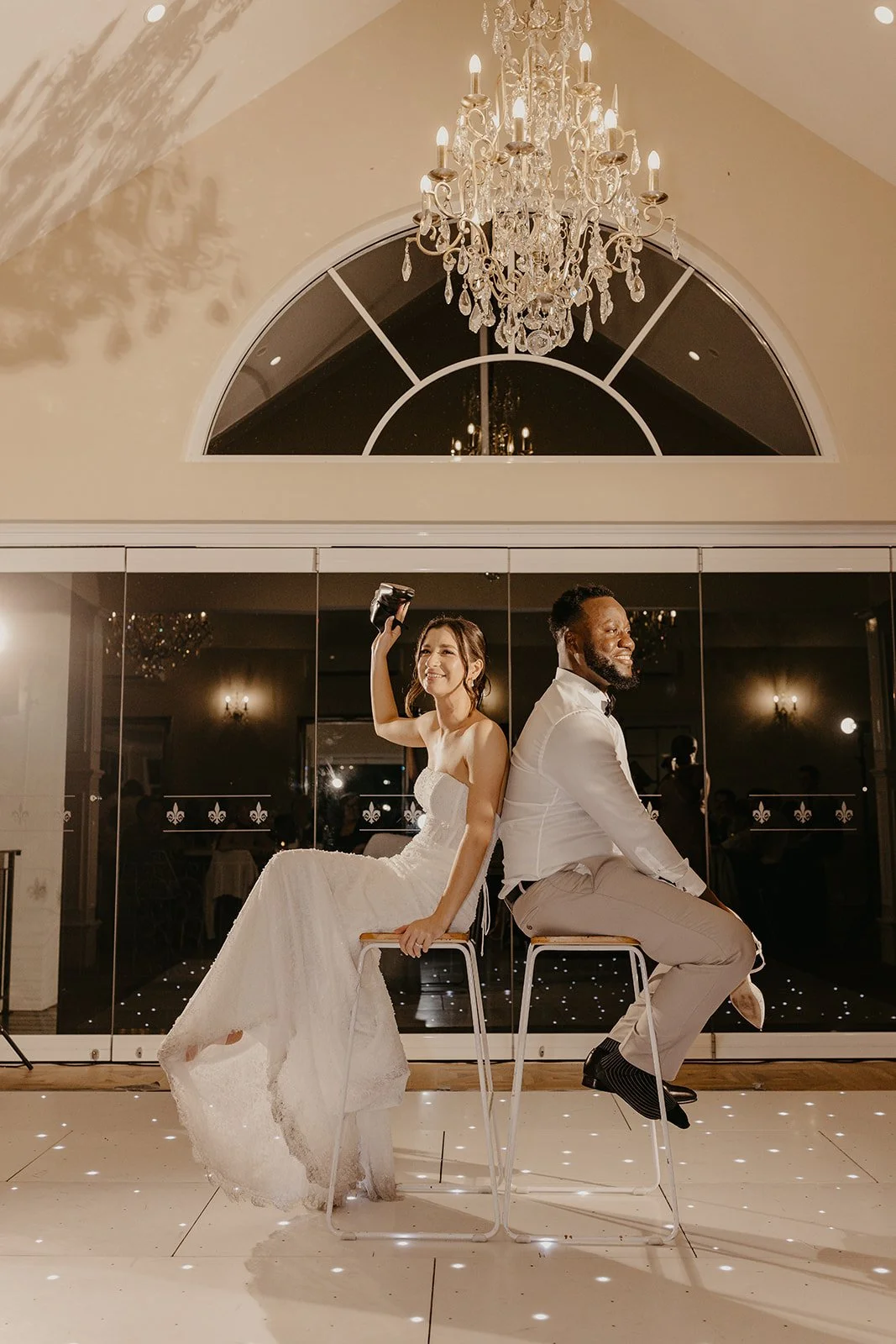 Bride and groom seated back-to-back on tall chairs during their wedding reception, with a chandelier overhead and a mirrored wall behind them.