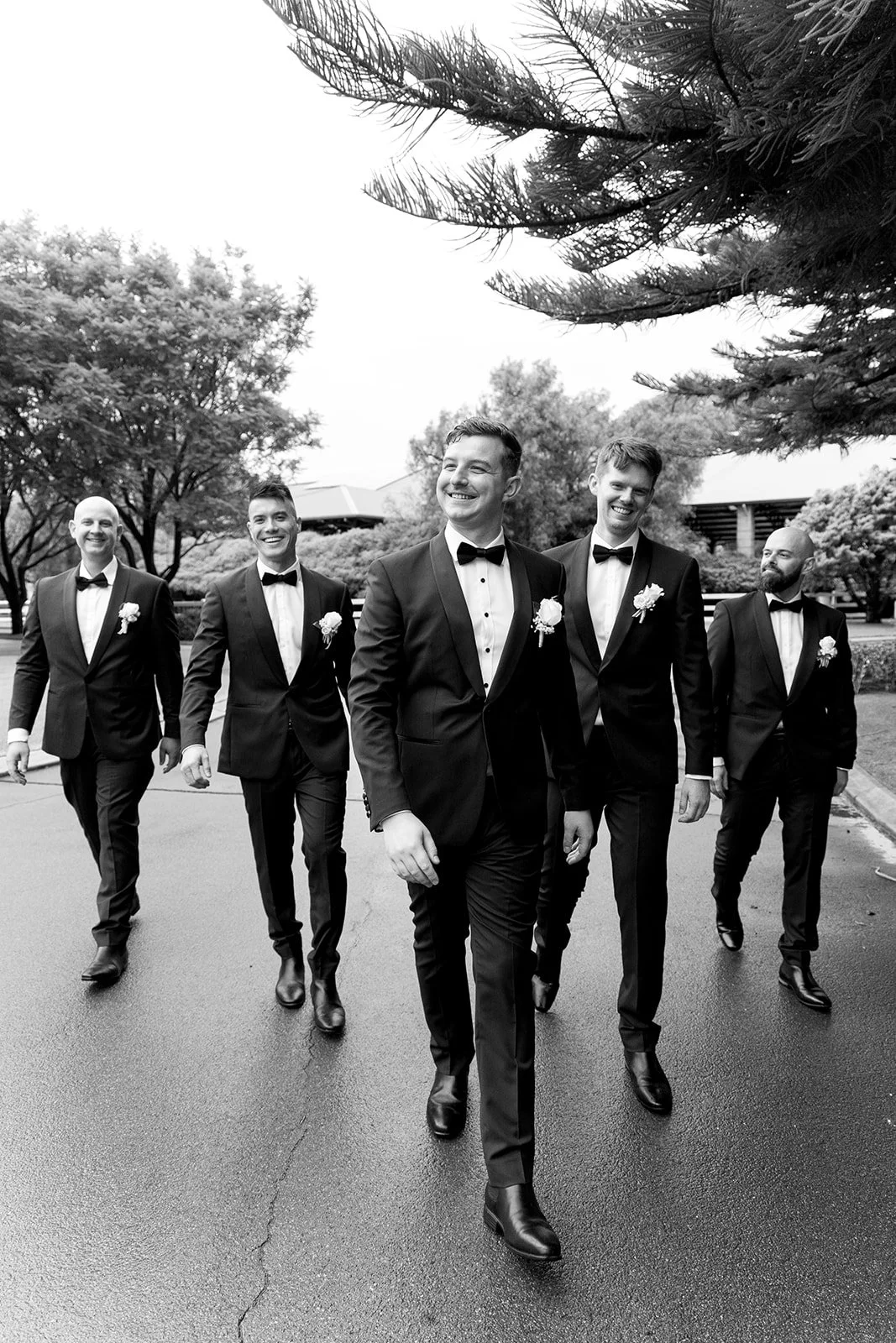 A group of six men walking outdoors in formal tuxedos with bow ties and boutonniers, smiling, with trees in the background.