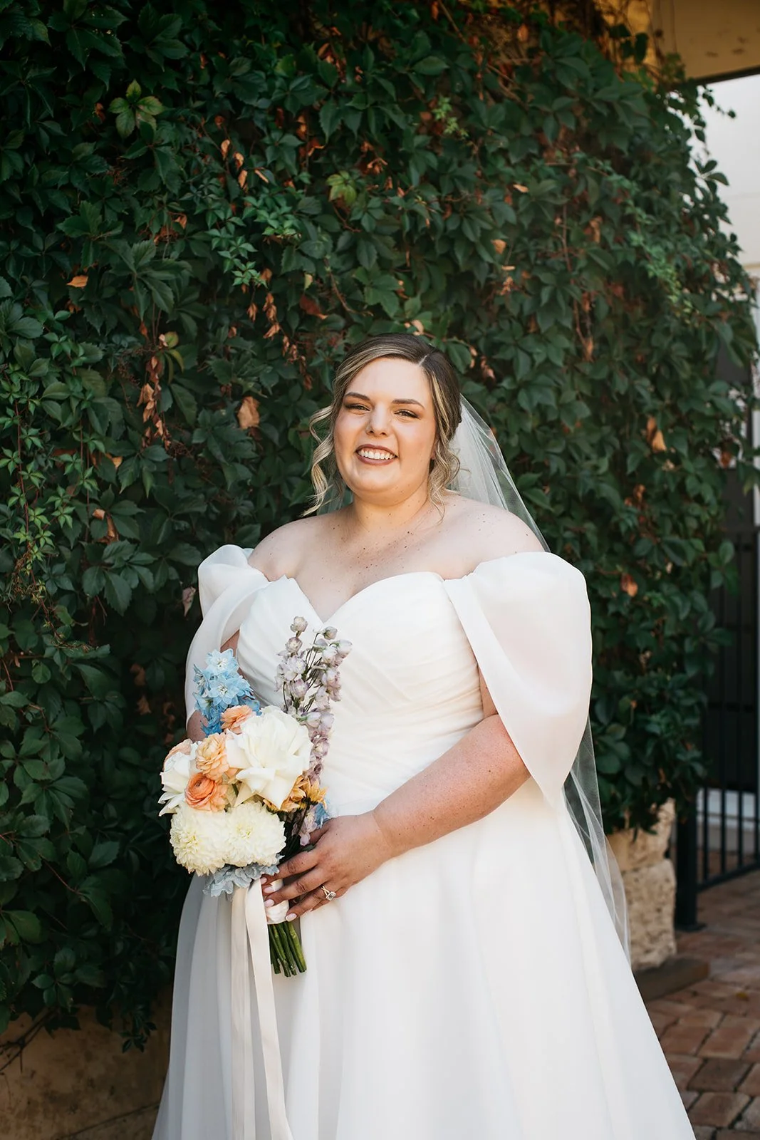A smiling bride in a wedding dress holding a bouquet of flowers, standing outdoors against a green leafy background.