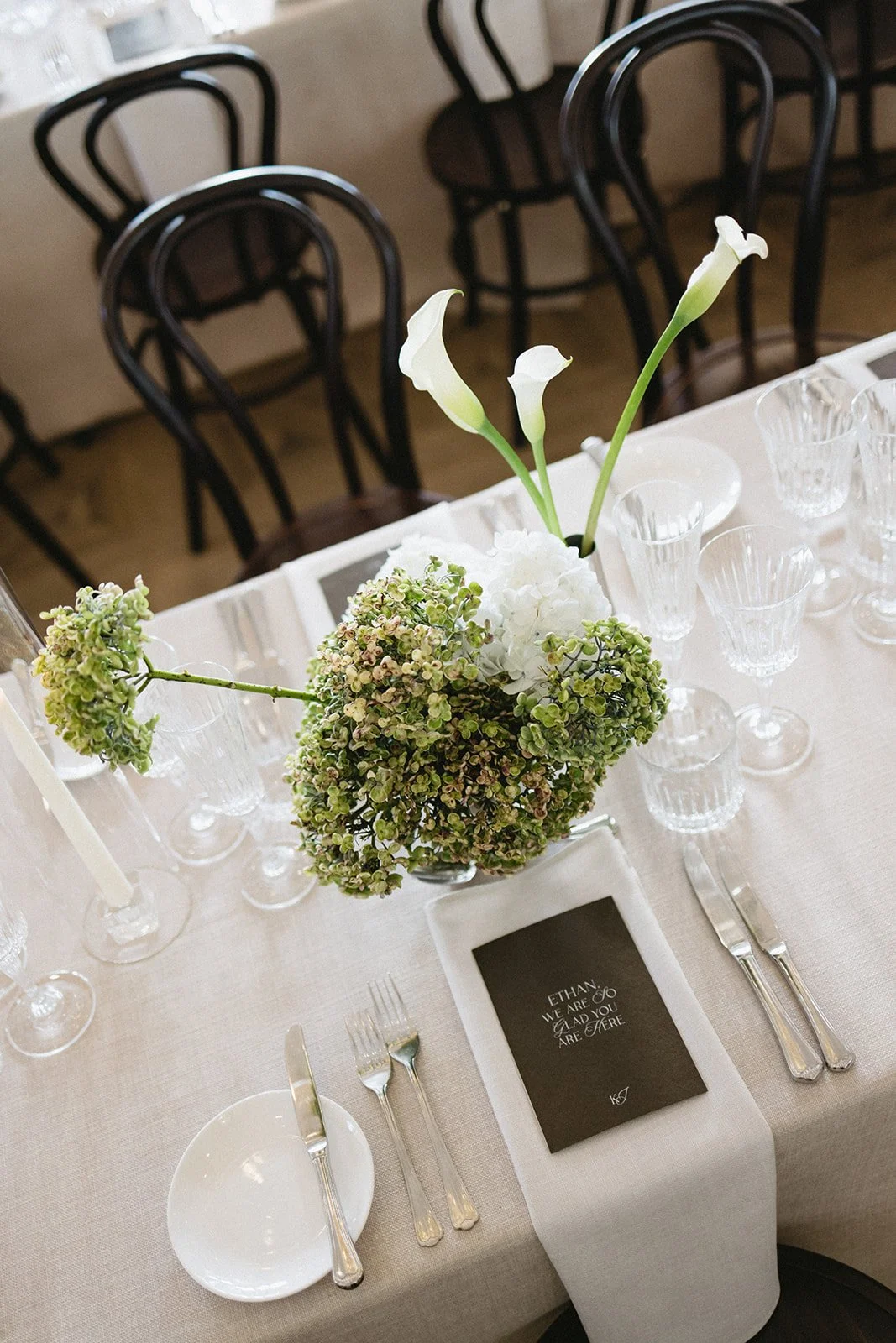 A floral centerpiece with white calla lilies and hydrangeas on a formal dining table set with glassware, silverware, and a small black paper with a closing message.