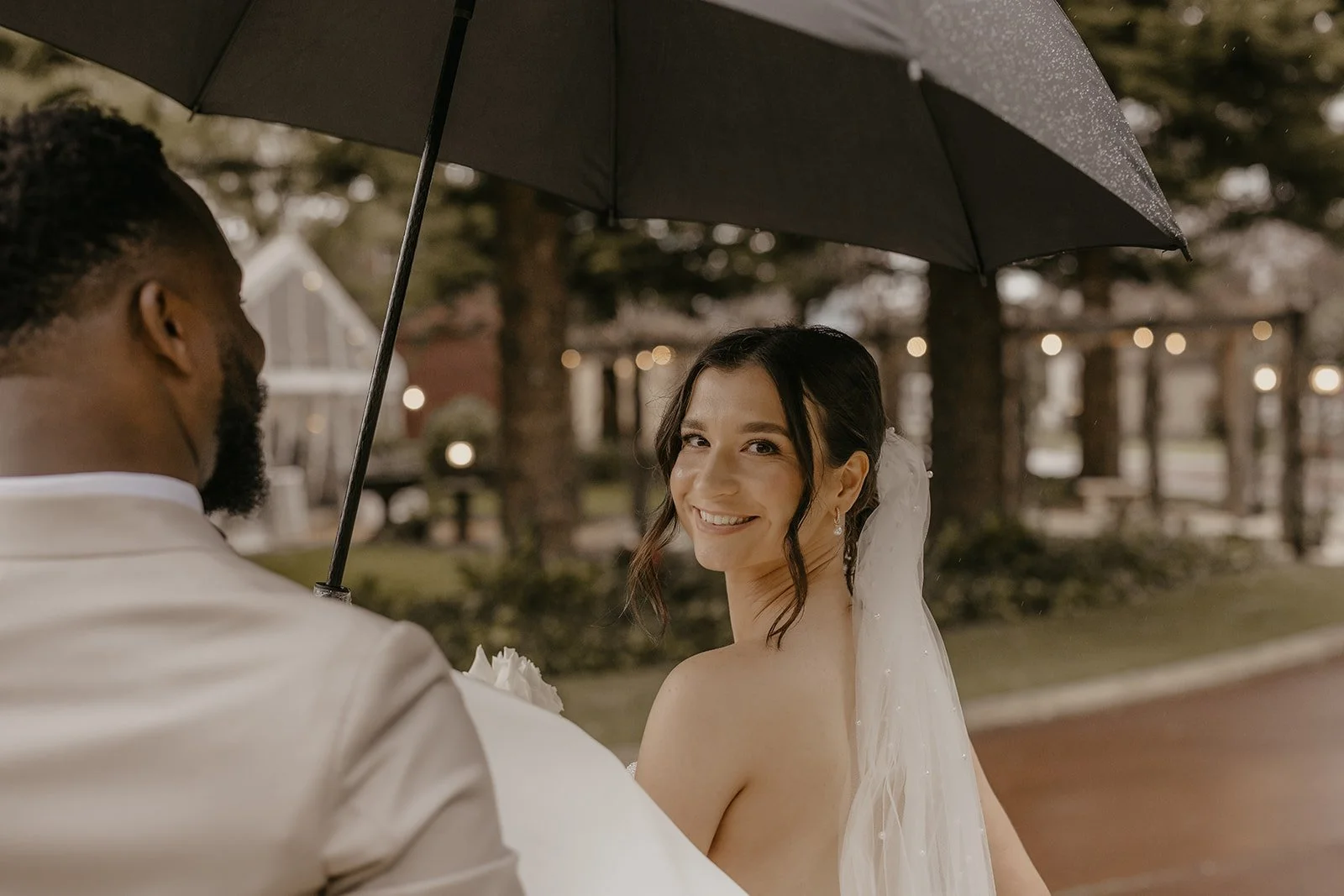 A bride with dark hair smiling and looking at the camera, holding a bouquet, standing under a black umbrella, with a groom facing her, outdoors in a park-like setting with trees and string lights in the background.