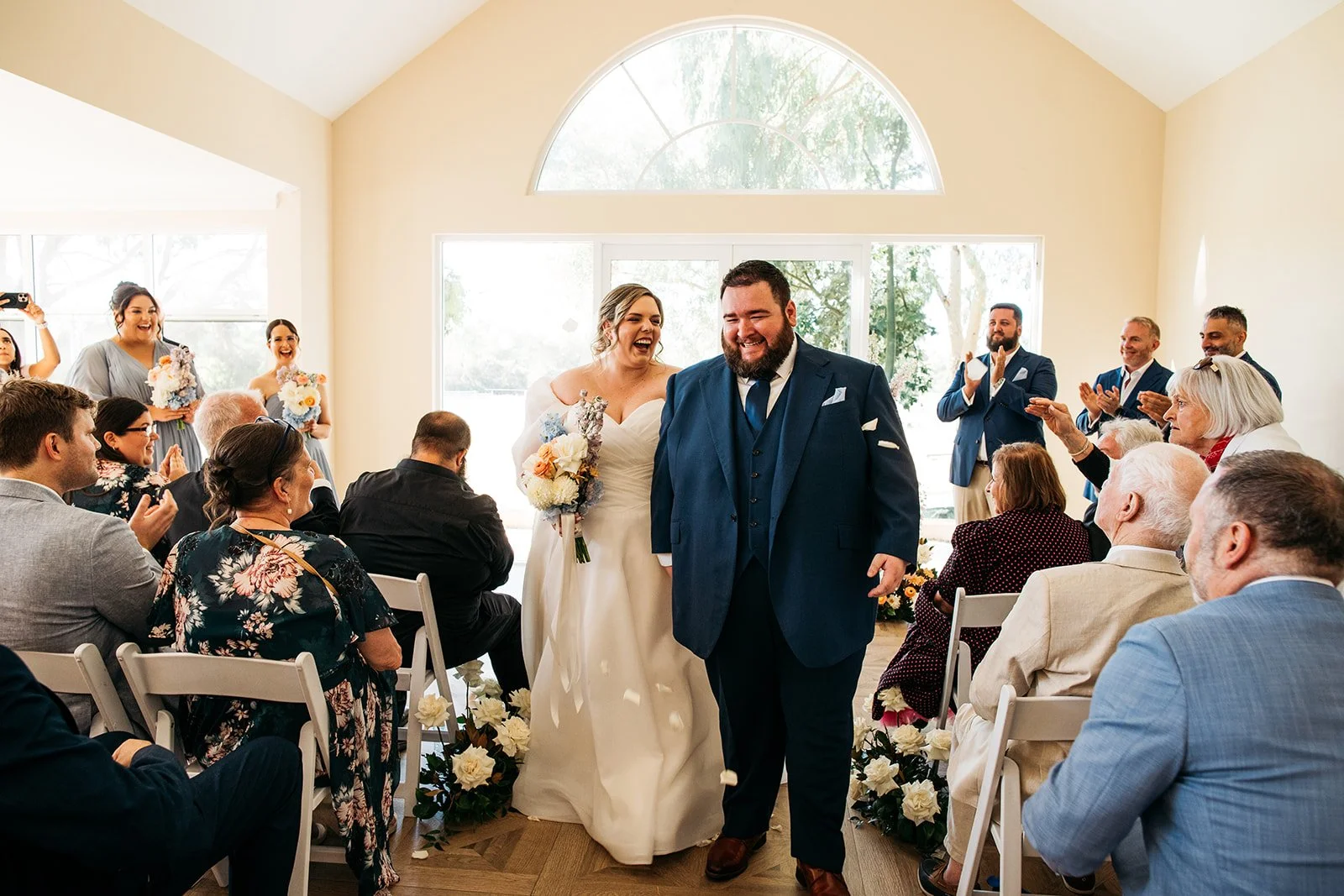 A bride and groom walking down the aisle during a wedding ceremony, smiling and sharing a joyful moment while surrounded by seated guests and bridesmaids and groomsmen clapping and celebrating.