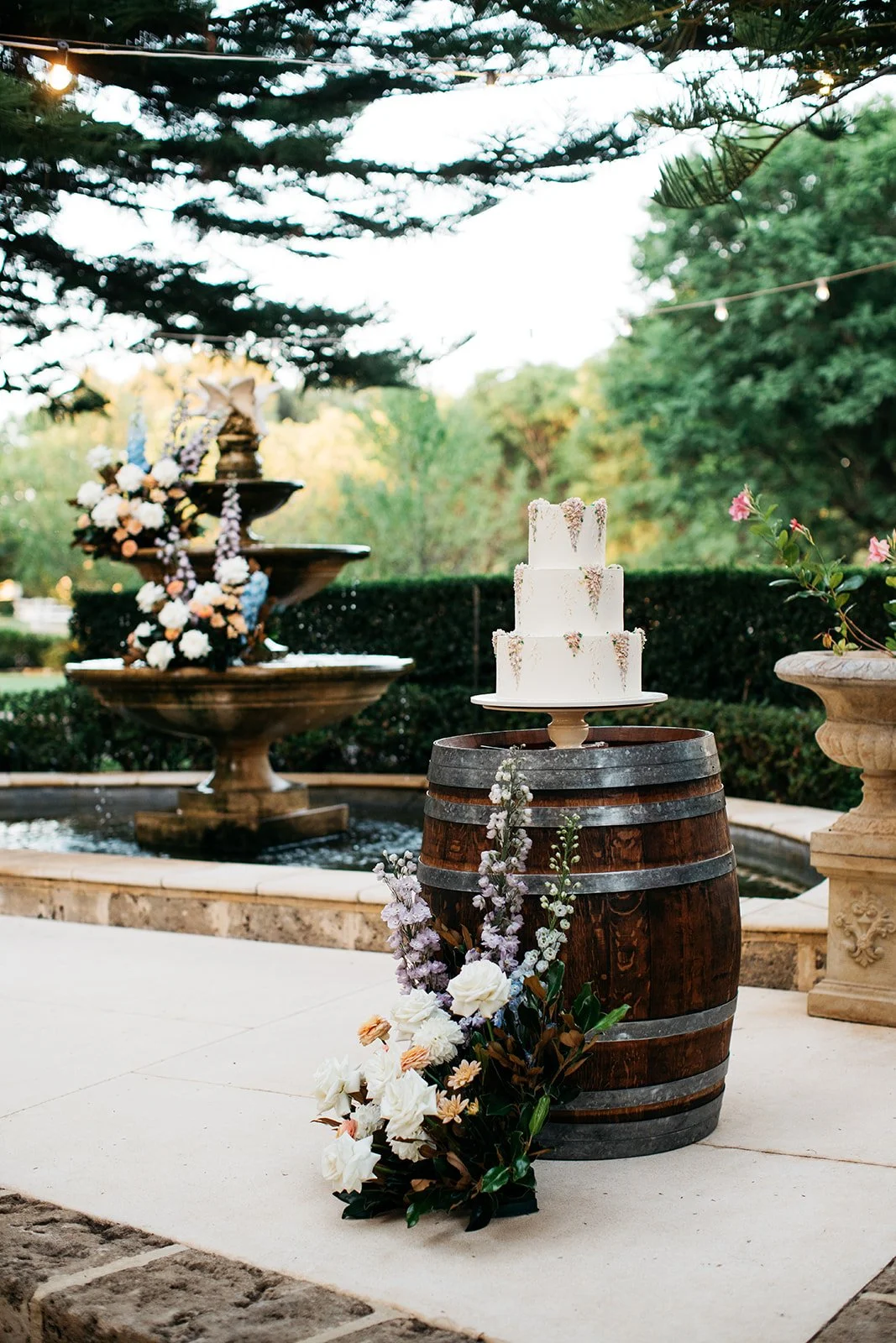 A three-tiered white wedding cake decorated with sprigs of flowers on a cake stand atop a wooden barrel, surrounded by flowers, with a fountain decorated with flowers in the background.
