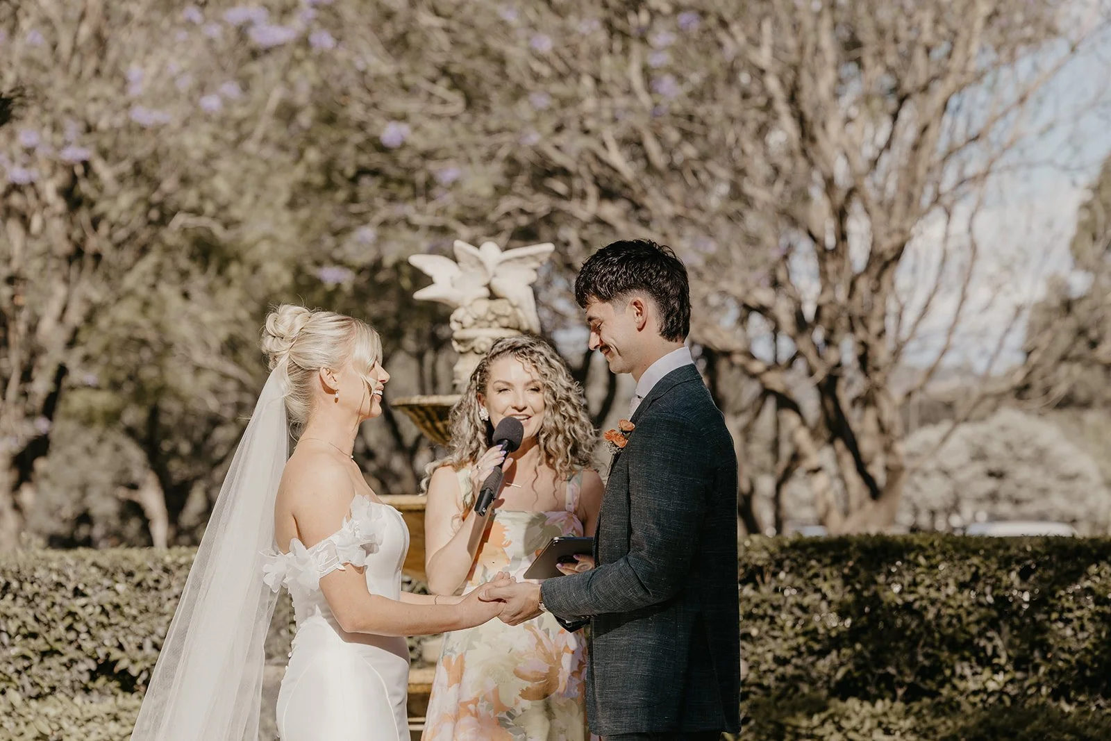A bride and groom holding hands during their outdoor wedding ceremony, with a female officiant speaking into a microphone, set against a background of trees and blue sky.