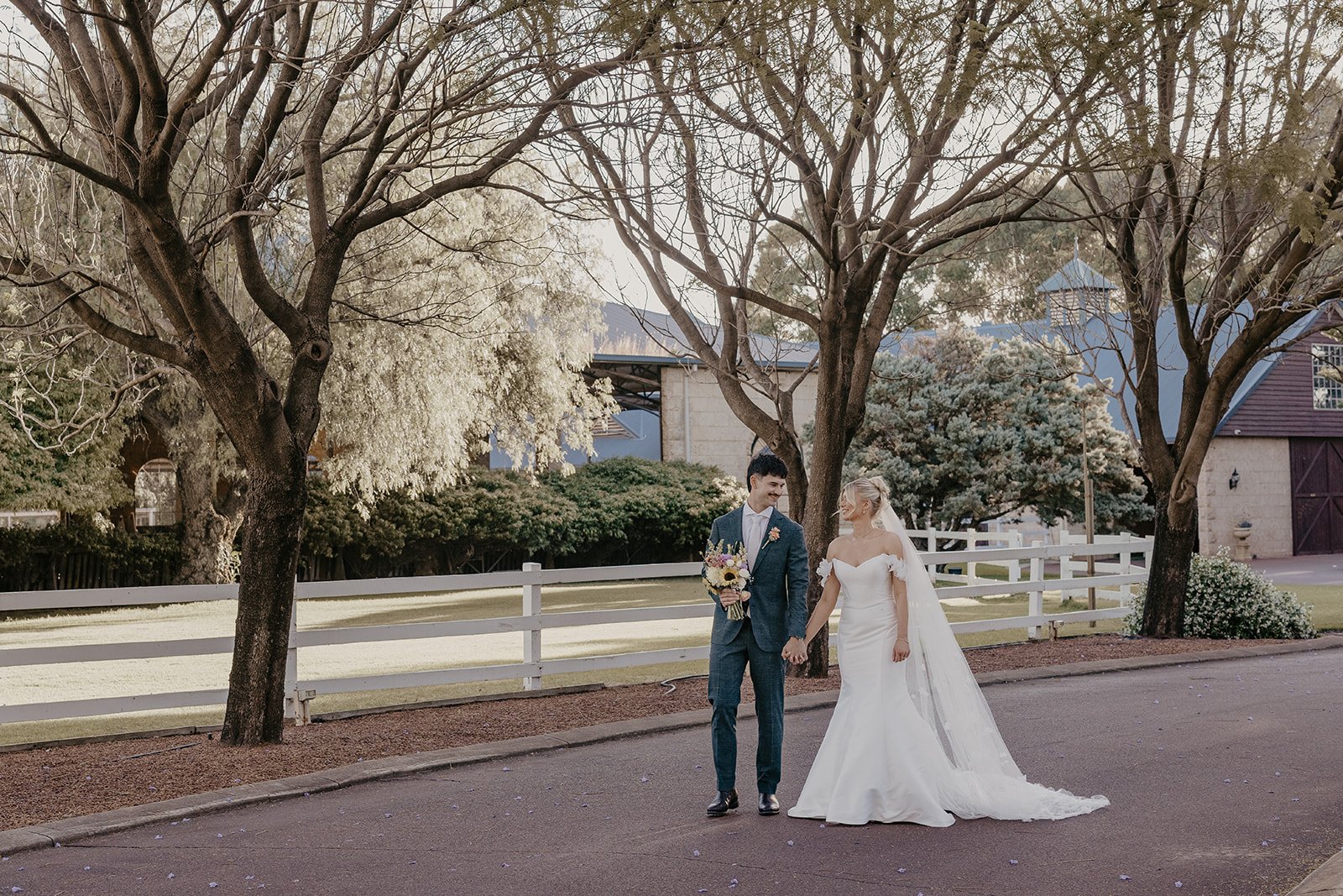 A bride and groom walking hand in hand outdoors on a wedding day, surrounded by trees and a white fence.