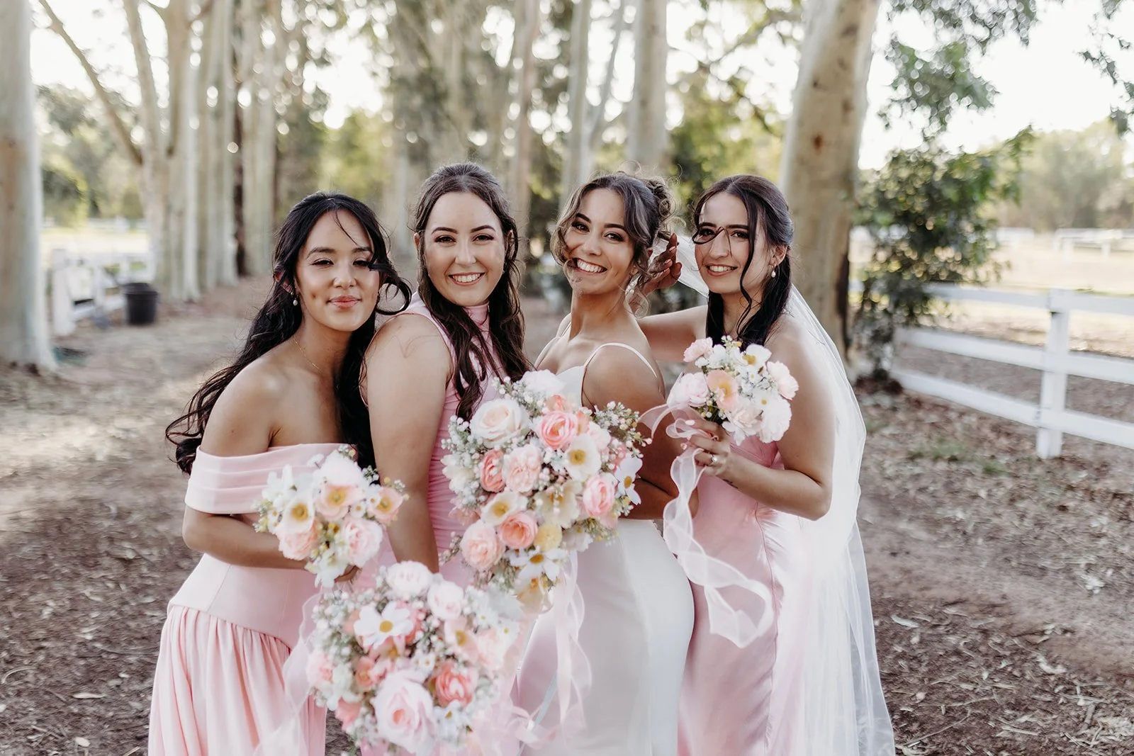 Four women in pink dresses holding bouquets of flowers, smiling, standing outdoors near trees and a white fence.