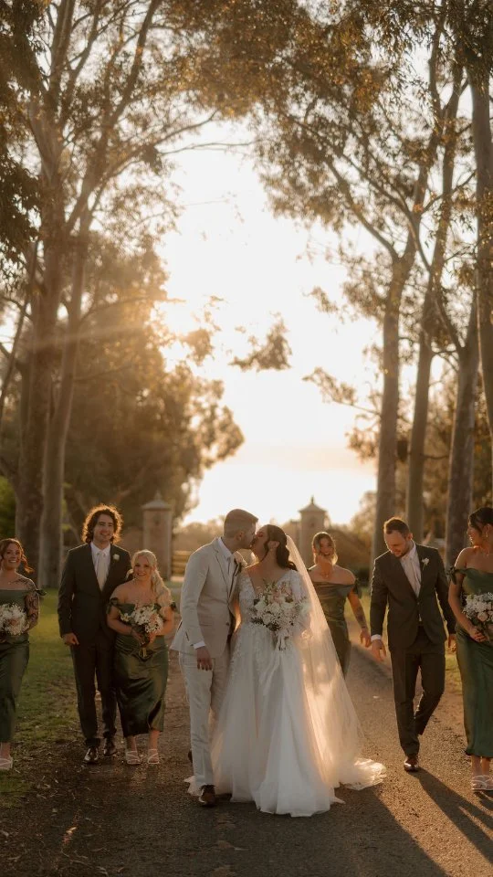 A wedding couple sharing a kiss while walking outdoors at sunset, surrounded by bridesmaids and groomsmen in formal attire.