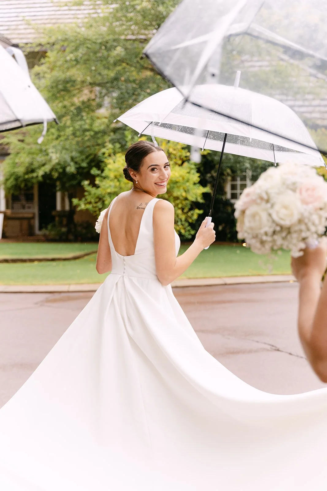 A smiling bride in a white wedding dress holding an umbrella outdoors, with a blurred bouquet in the foreground and greenery in the background.