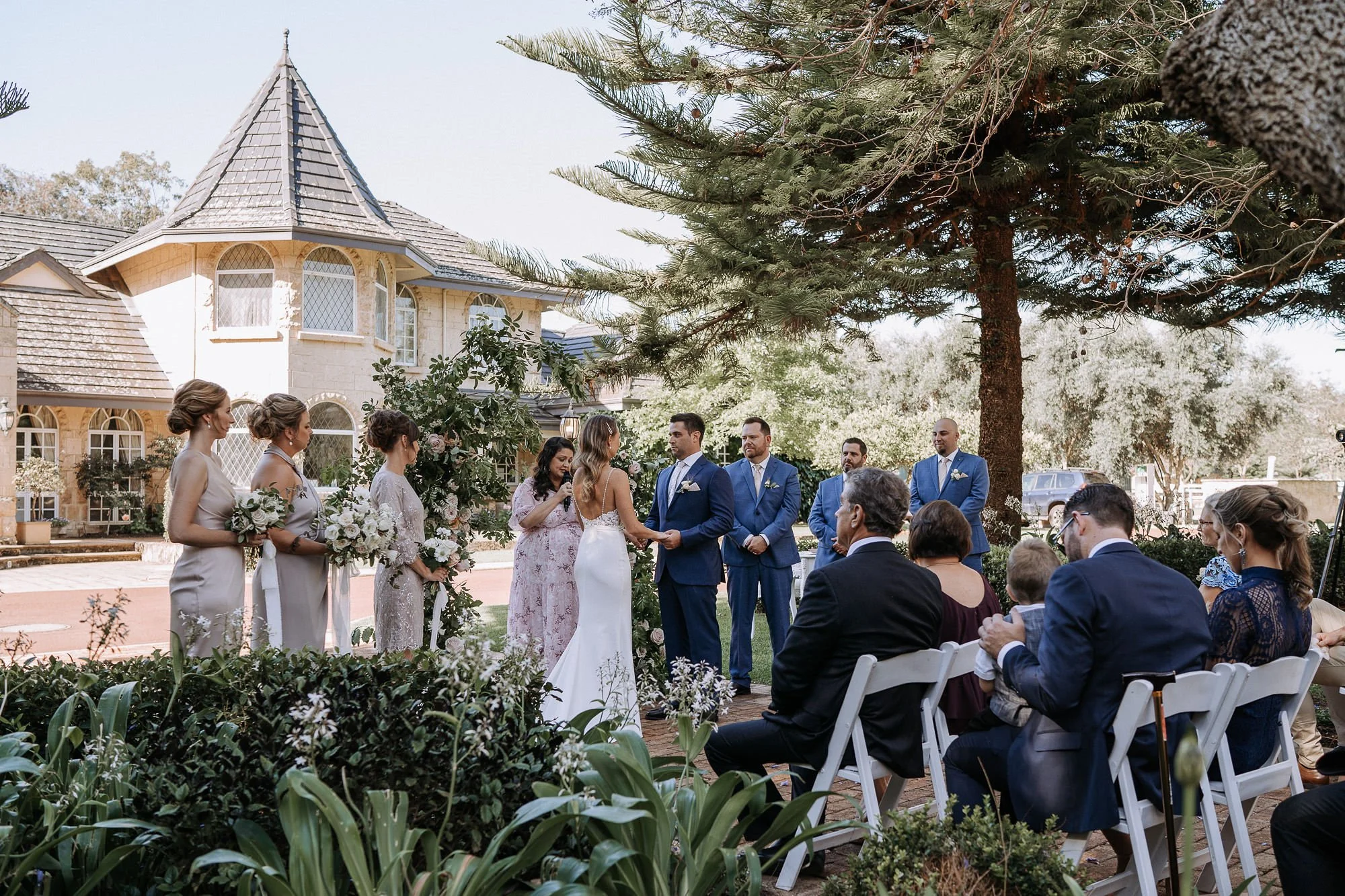 A wedding ceremony outdoors with the bride and groom standing hand-in-hand, surrounded by bridesmaids and groomsmen, in front of a large tree and elegant house, with seated guests watching.
