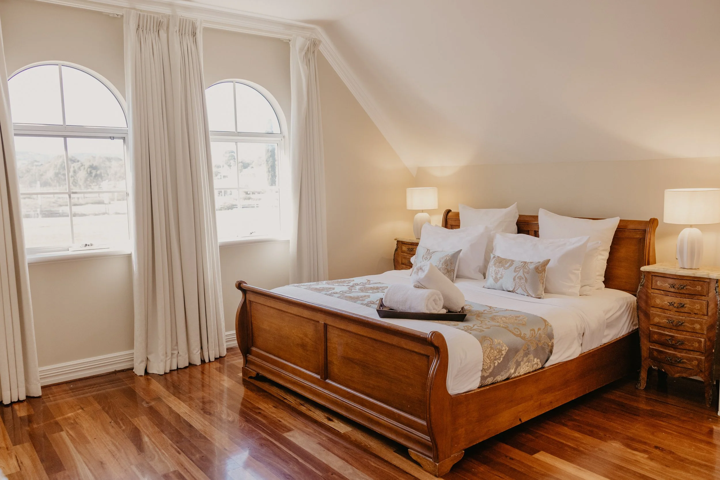 A bedroom with a large wooden bed, white pillows, and a patterned bedspread. There are two nightstands with lamps on each side of the bed. The room has three large arched windows with white curtains, and hardwood flooring.