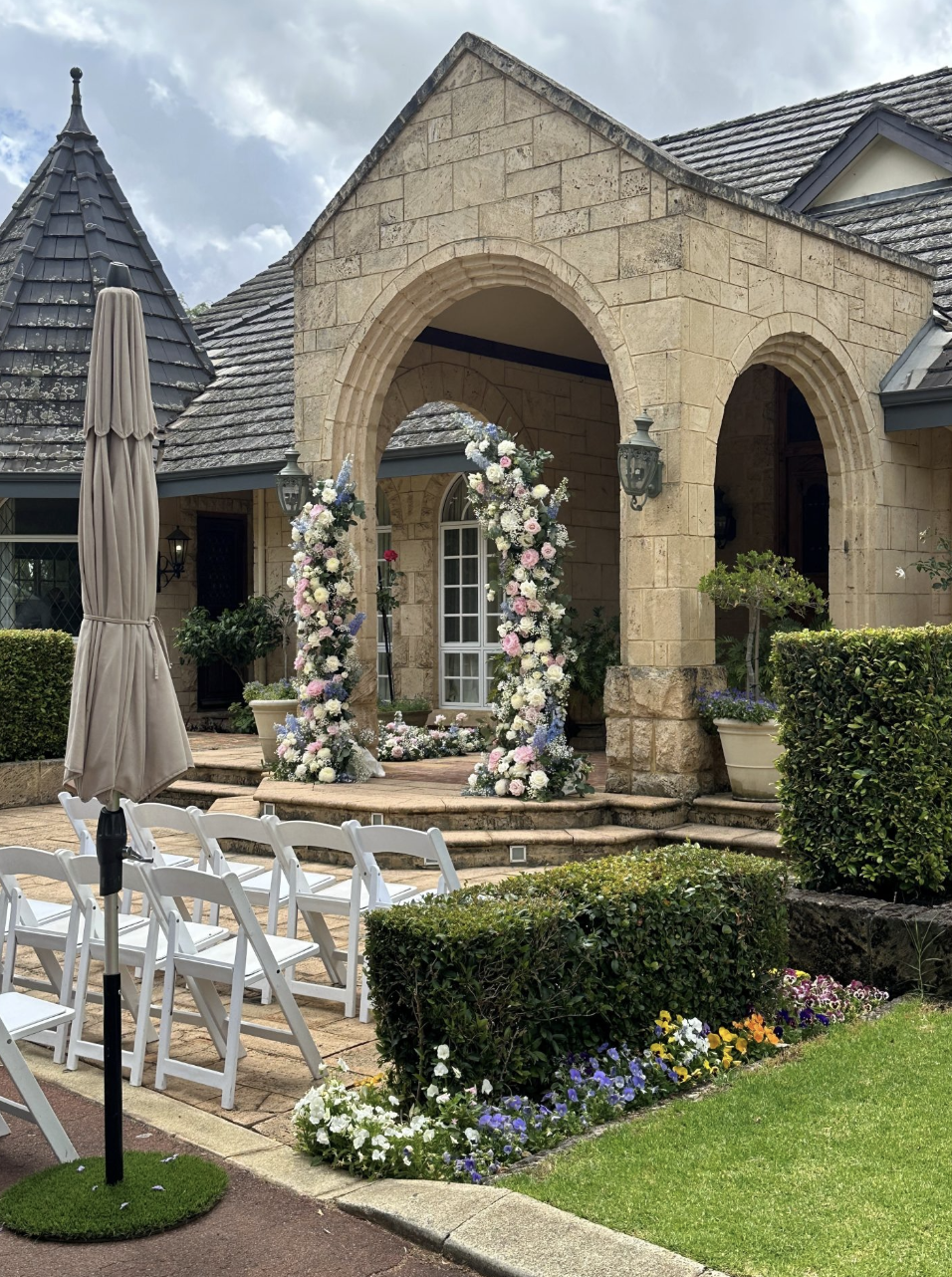 A wedding setup in front of a stone house with an arch decorated with flowers, white chairs arranged for guests, and a closed umbrella on the left.