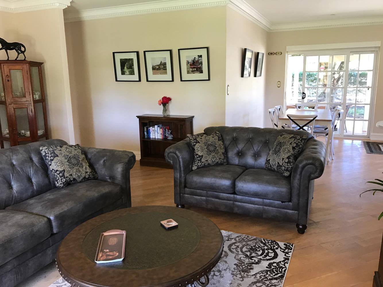 Living room with dark leather sofas, a round coffee table with a remote and a box, a small wooden bookshelf, a corner display cabinet, framed pictures on the wall, a dining table with chairs near a sliding door leading outside, and hardwood floors.