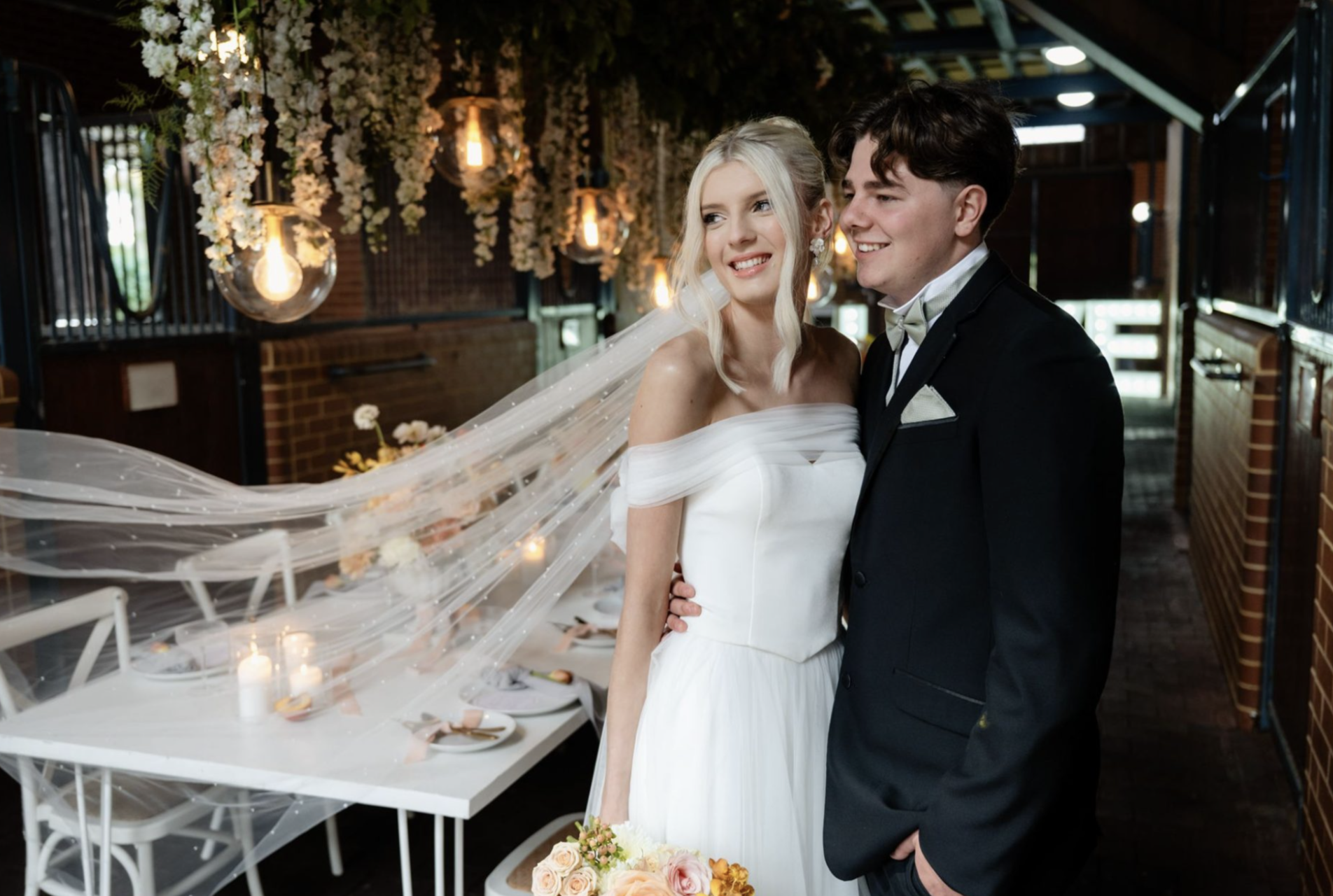 A bride and groom are smiling and standing close together at their wedding reception indoors, decorated with hanging white flowers and warm lighting.