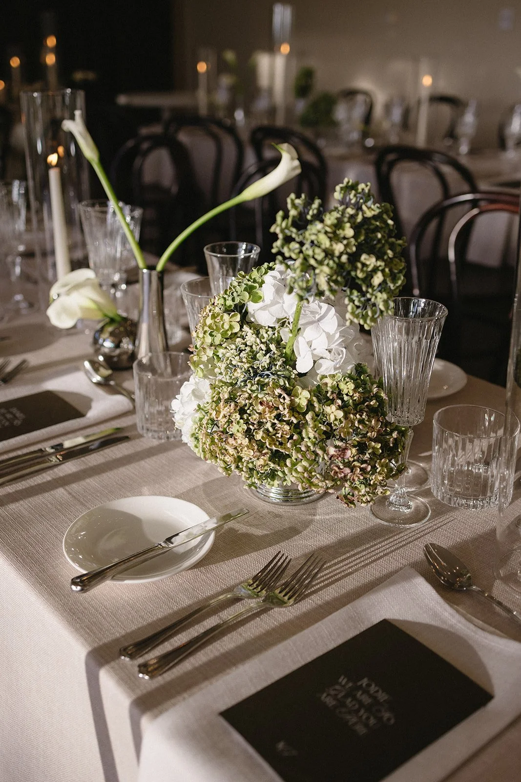 Elegant dining table decorated with a floral centerpiece consisting of white and green flowers, including hydrangeas and calla lilies, surrounded by glassware, cutlery, black menus, and a white tablecloth in a dimly lit setting.
