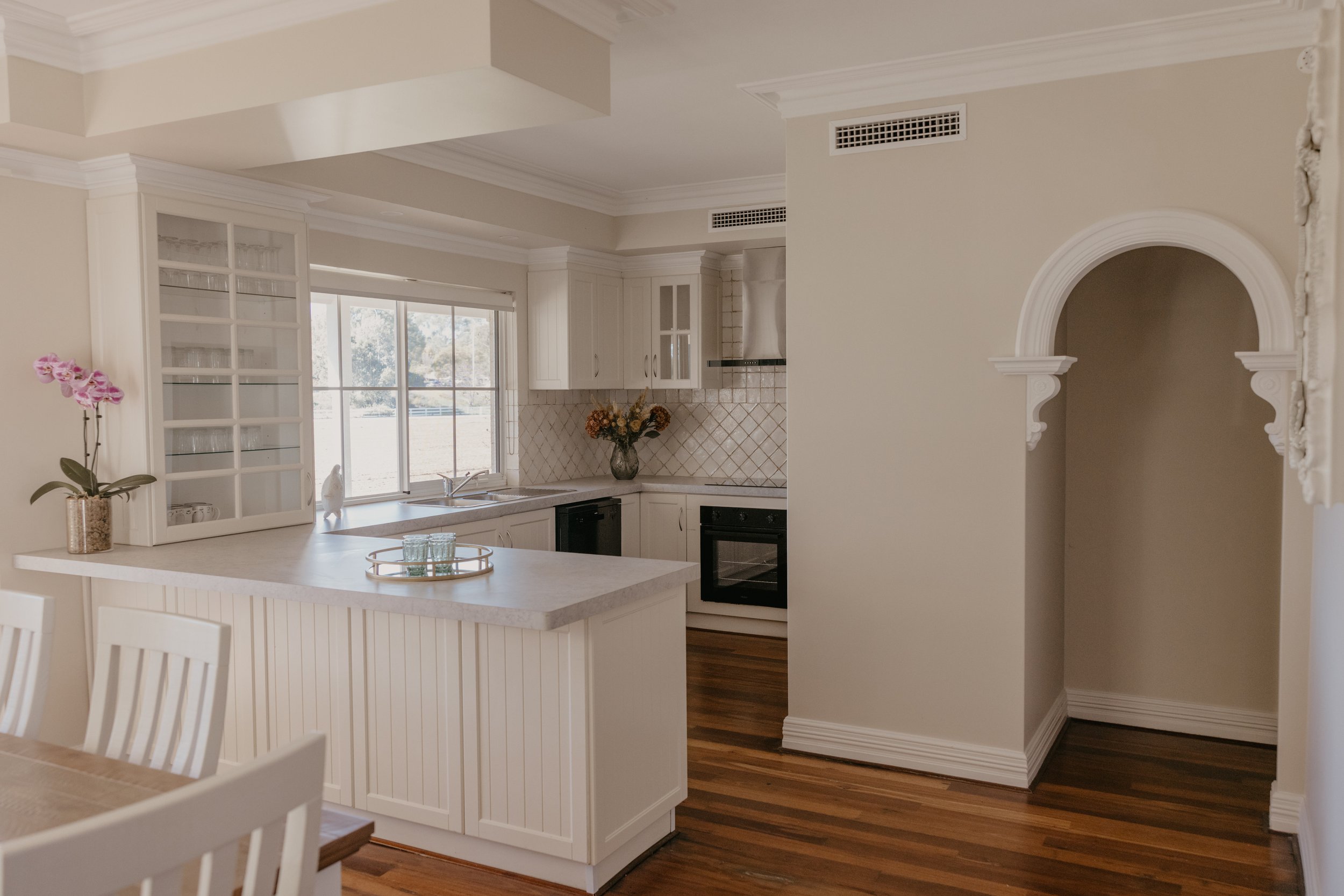 A kitchen with white cabinets, a window with natural light, a beige floral vase with flowers, a white kitchen island, and a dining table with white chairs. There is a decorative archway on the right side.