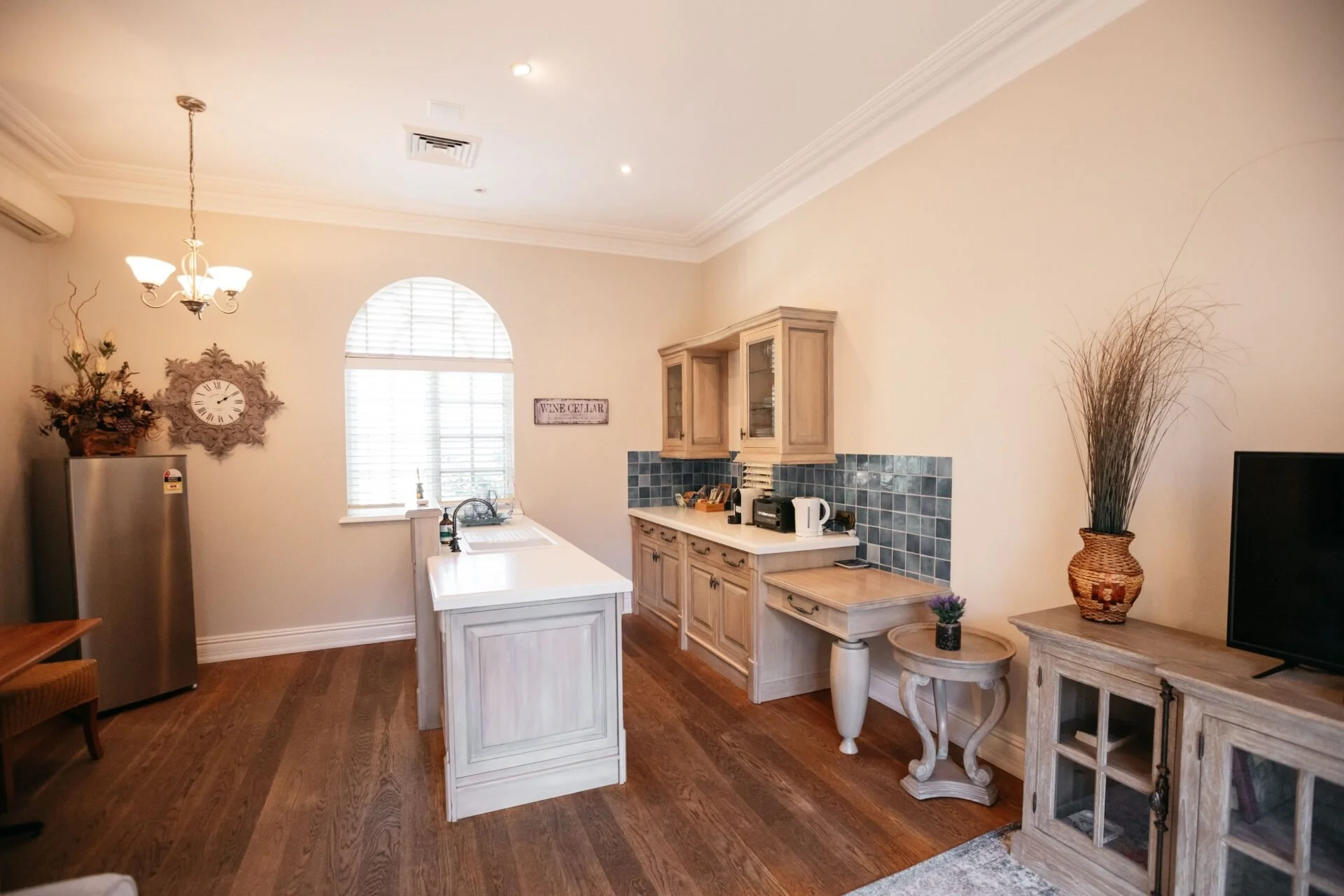 A cozy kitchen with white cabinetry, a small island, and wooden flooring. Features include a window, a decorative wall clock, and various small appliances on the counters. A large wicker vase with tall dried grass is on the right side of the image.