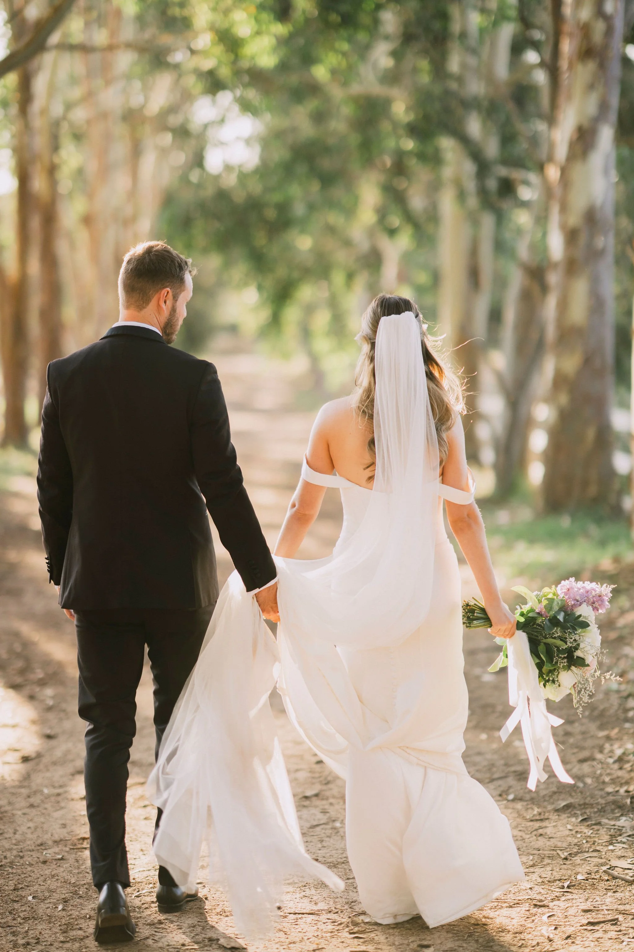A bride and groom walking hand in hand through a forested area during wedding, sunlight filtering through trees, bride holding a bouquet.