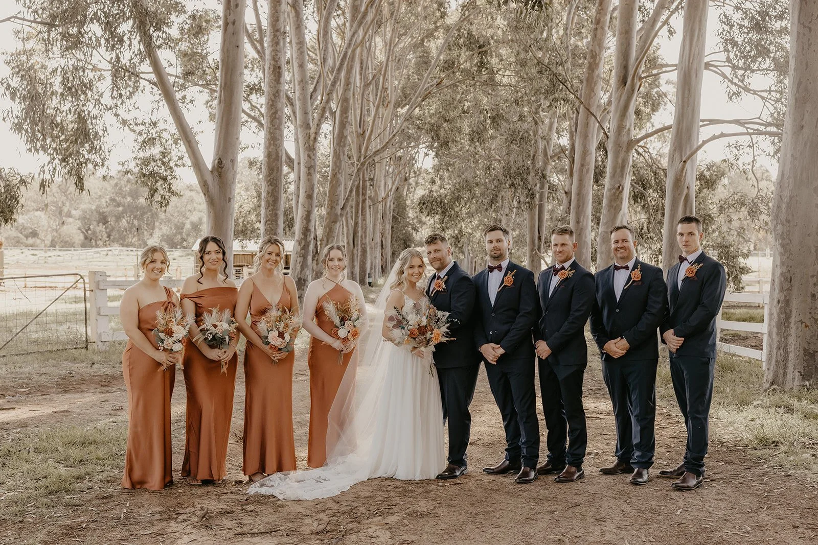 A wedding party with five bridesmaids in rust-colored dresses and five groomsmen in dark suits with bow ties, standing outdoors among tall trees in what appears to be a rustic setting.