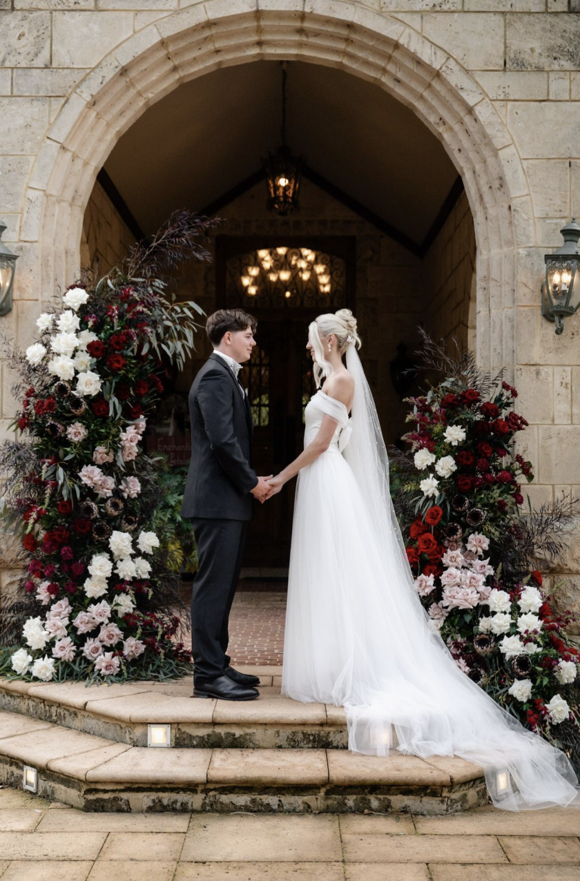 A bride and groom holding hands and facing each other under a stone archway, decorated with red, white, and pink flowers, at their wedding ceremony outside a stone building with lanterns.