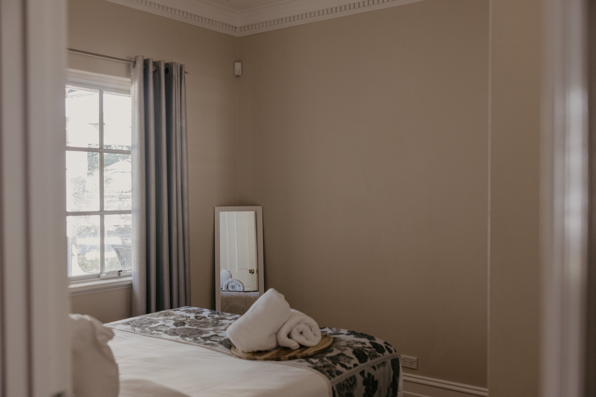 A bedroom seen through a partially open door, featuring a window with gray curtains, a mirror, a bed with a floral bedspread, and rolled towels on the bed.