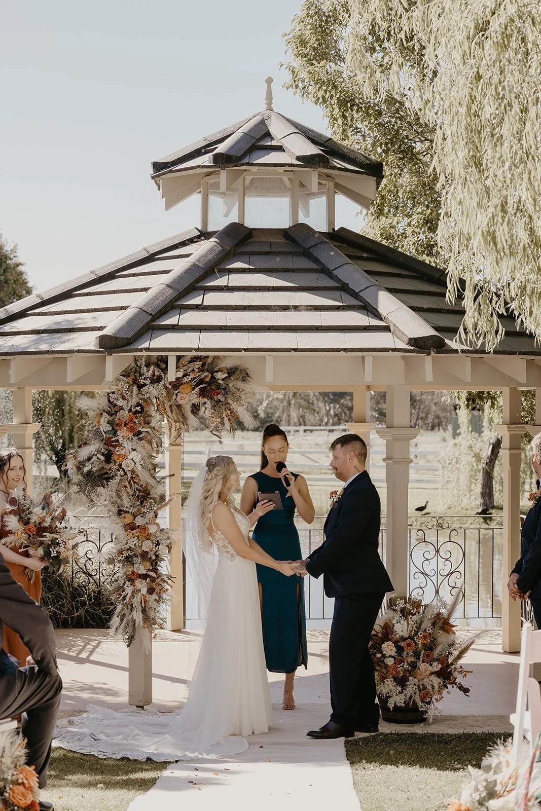 A couple getting married at Brookleigh Estate holding hands during their wedding ceremony in an outdoor gazebo decorated with flowers and foliage, with an officiant and guests present.