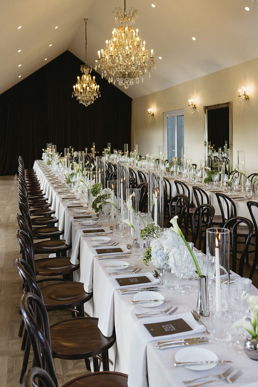 Elegant wedding reception with long white-draped tables, dark wooden chairs, tall glass centerpieces with white flowers, and hanging chandeliers in a spacious room.