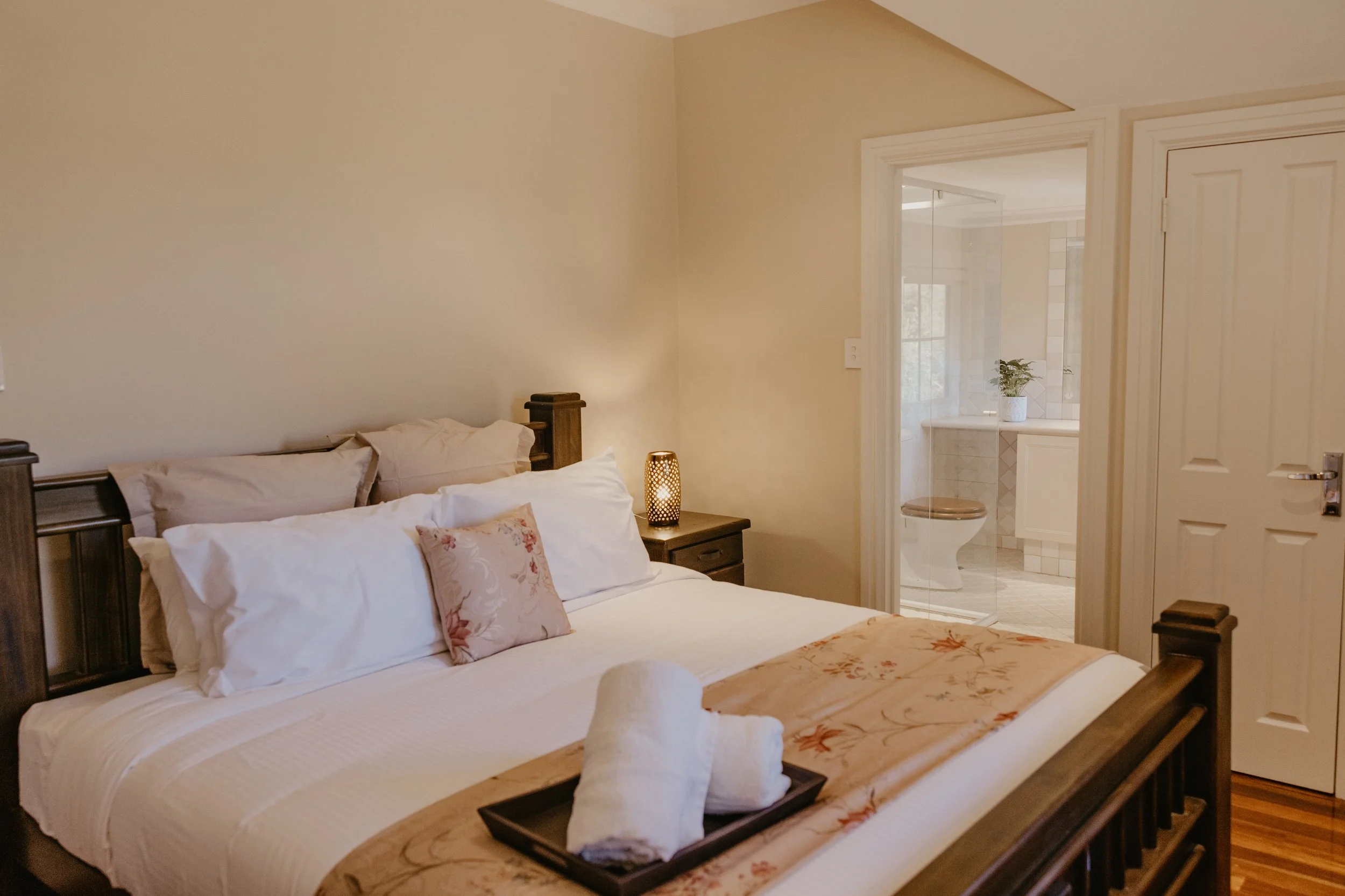 A bedroom with a bed, pillows, and a blanket, with a nightstand that has a lamp. There is a door leading to a bathroom with a visible toilet and a window with a potted plant.