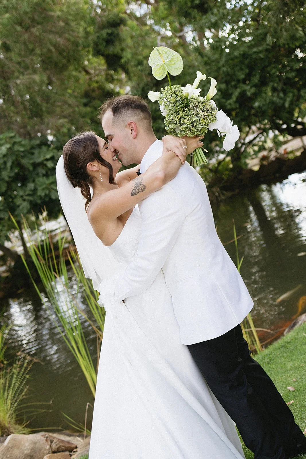 A newlywed couple embracing outdoors by a pond, with green trees in the background. The bride is holding a bouquet and wearing a white wedding dress, and the groom in a white suit jacket and black pants.