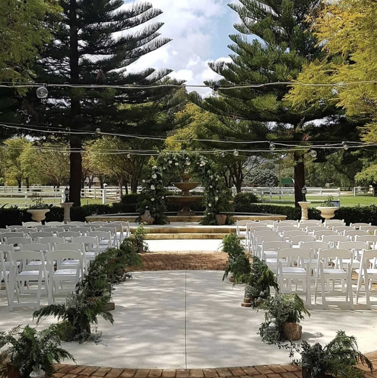 Outdoor wedding ceremony setup with white chairs arranged in rows facing a decorated altar with flowers and greenery, set in a park with large trees, a brick aisle, and a fountain in the background.