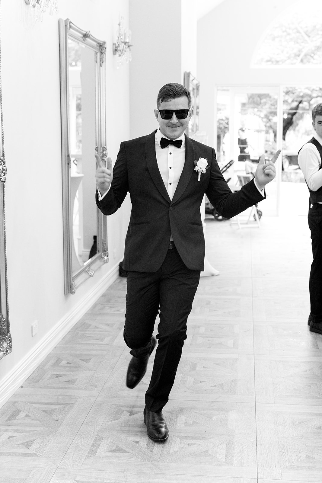 A man in a tuxedo, sunglasses, and a boutonnière is dancing and smiling at a formal event with mirrors and large windows in the background.
