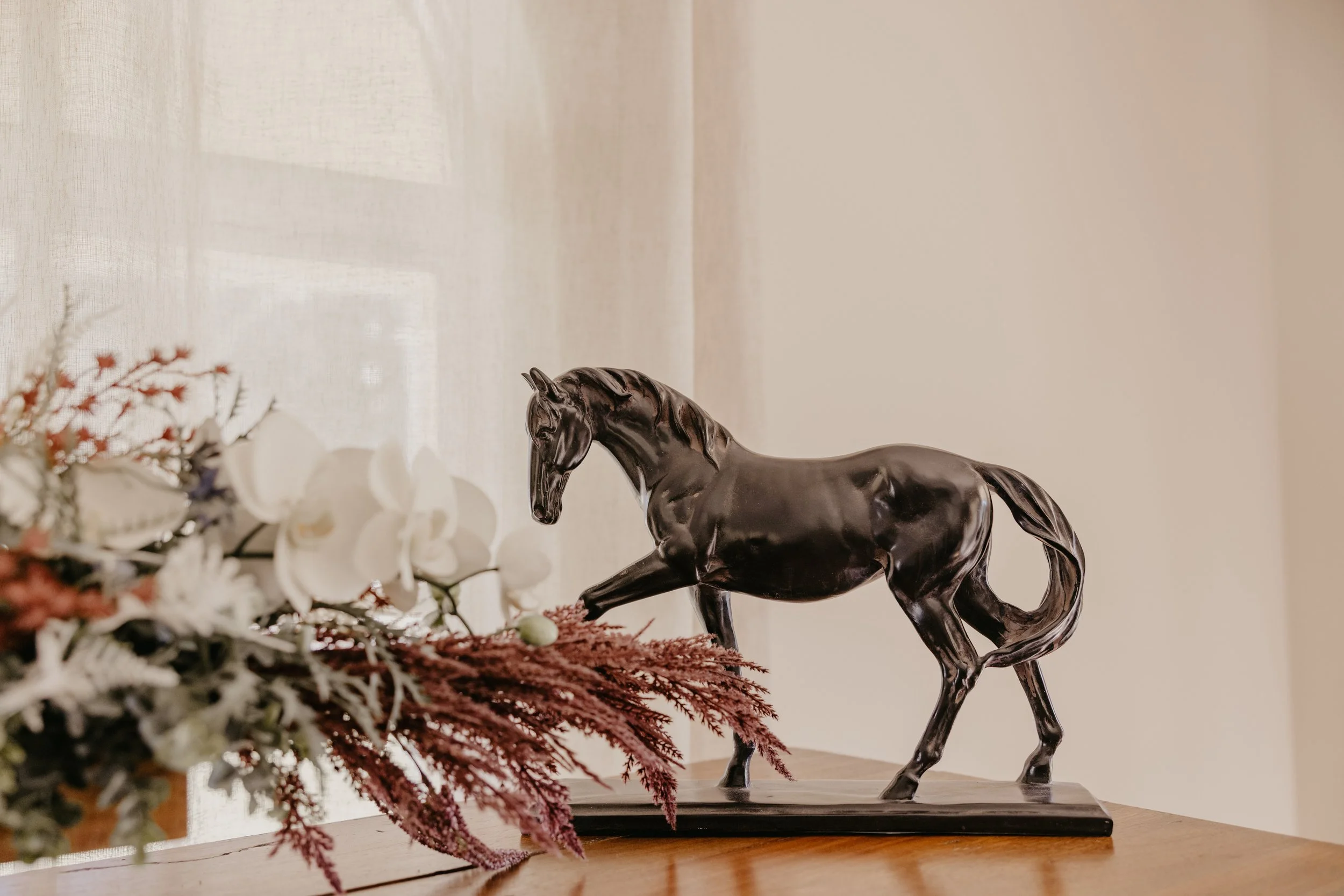 A decorative black metal horse sculpture standing on a rectangular base on a wooden surface, with a bouquet of flowers partly visible in the foreground and a beige-colored wall with a window covered by a sheer curtain in the background.