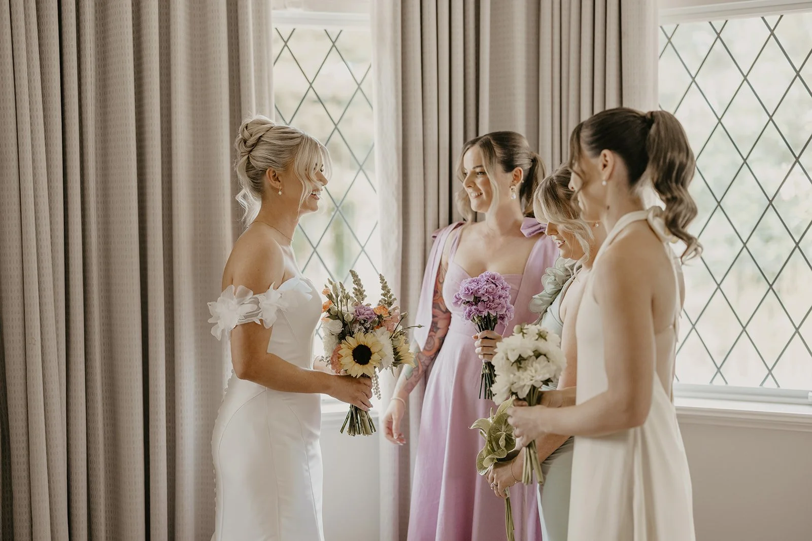 A bride with blonde hair in an updo, wearing a white off-shoulder wedding dress, holding a bouquet of sunflowers, roses, and greenery, smiling as she faces four women in pastel-colored dresses holding bouquets, in a room with large windows and beige 